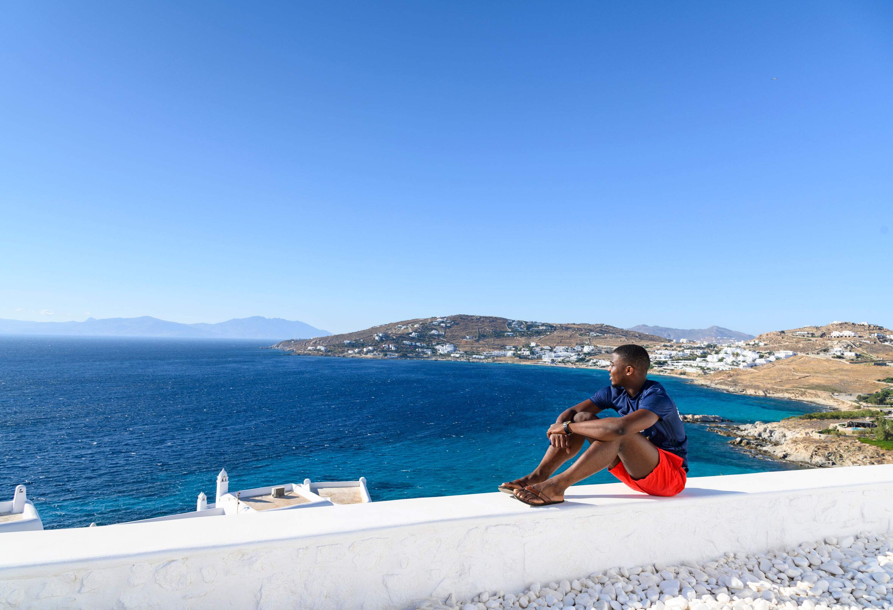 A man sits on a white wall looking at the tranquil blue sea against the clear blue sky.
