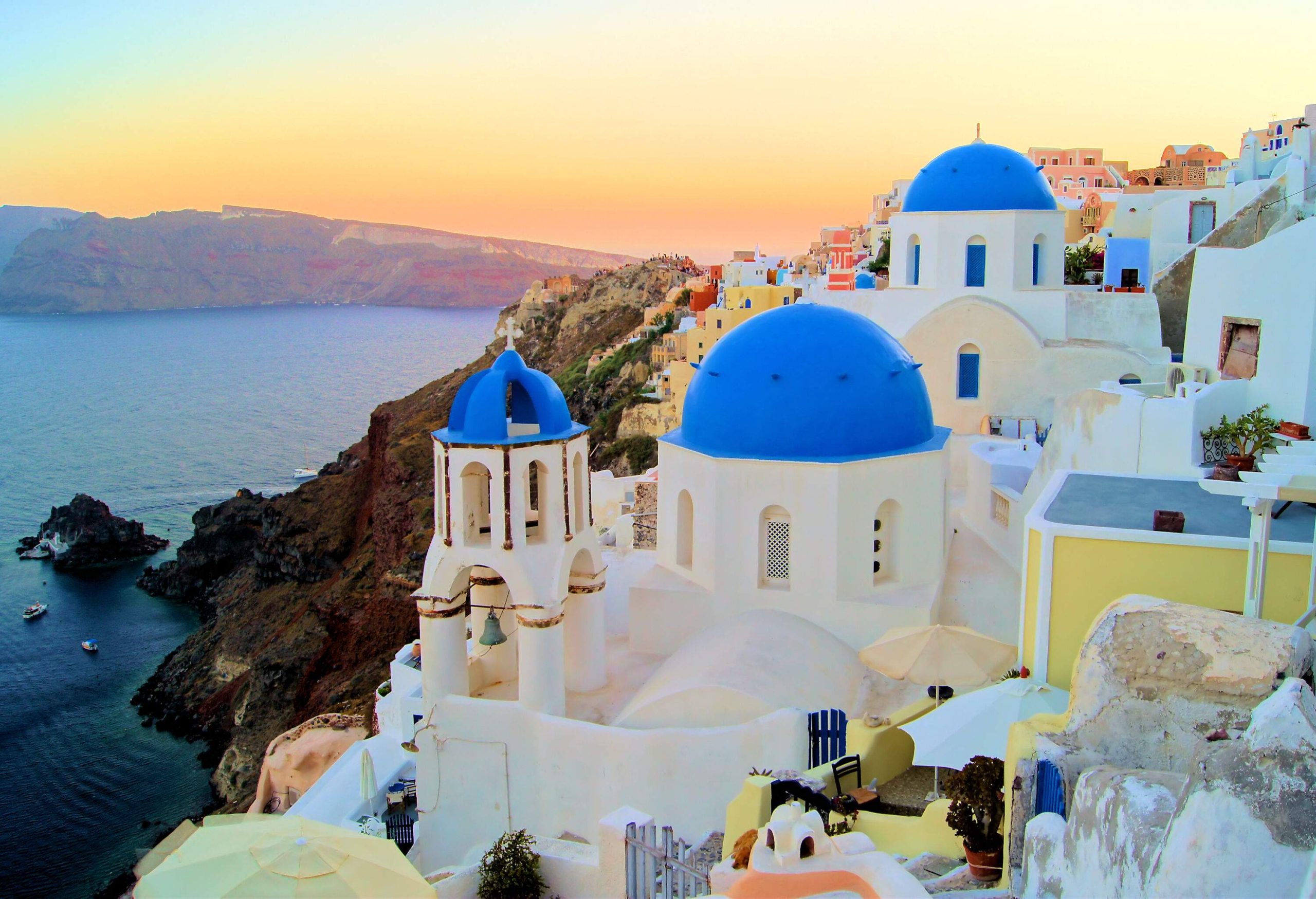 A blue dome church with a belfry in a hillside village set on a cliff's edge under an orange sky.