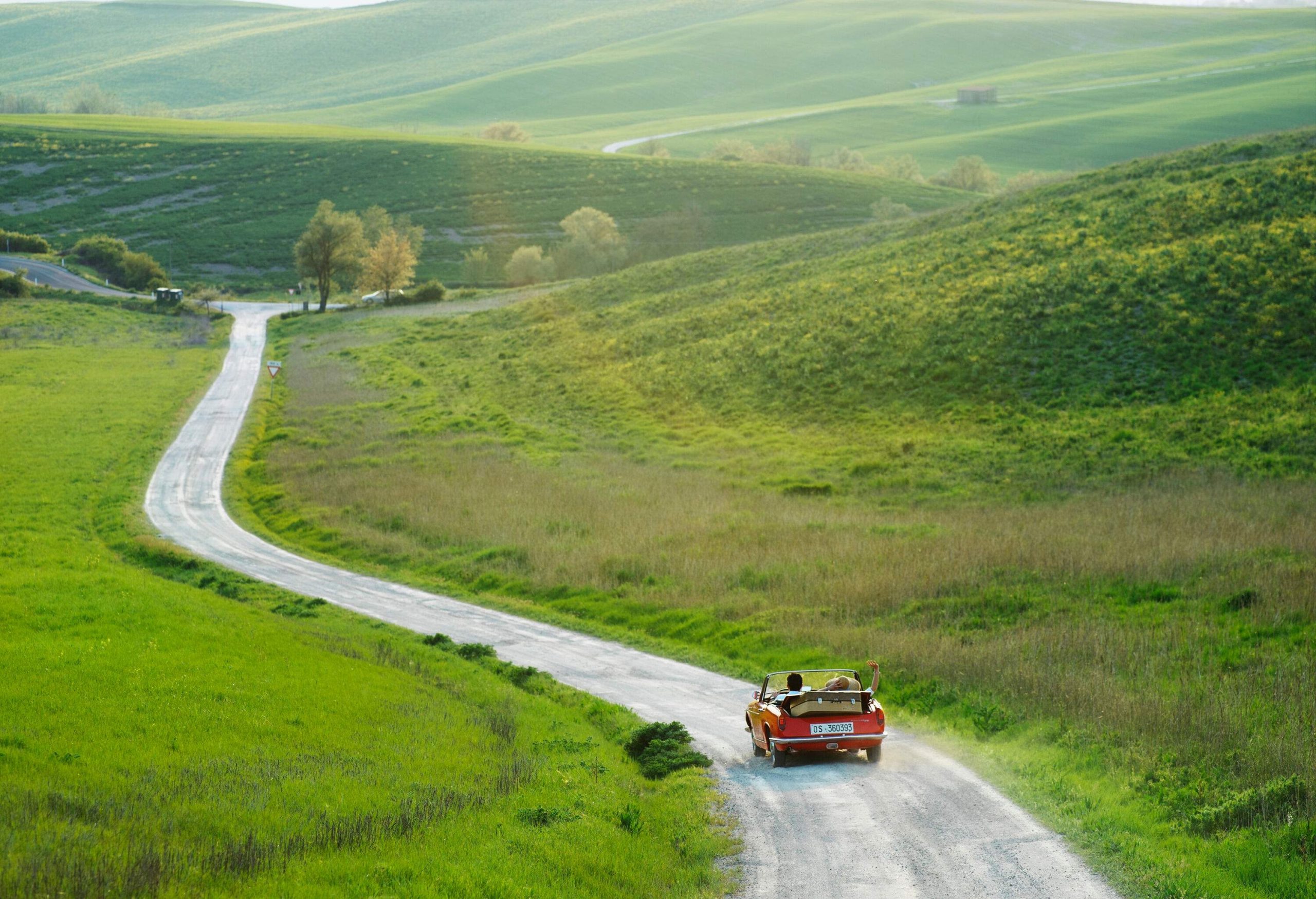 A couple in a red car travelling on an unpaved white downhill road towards the green fields.