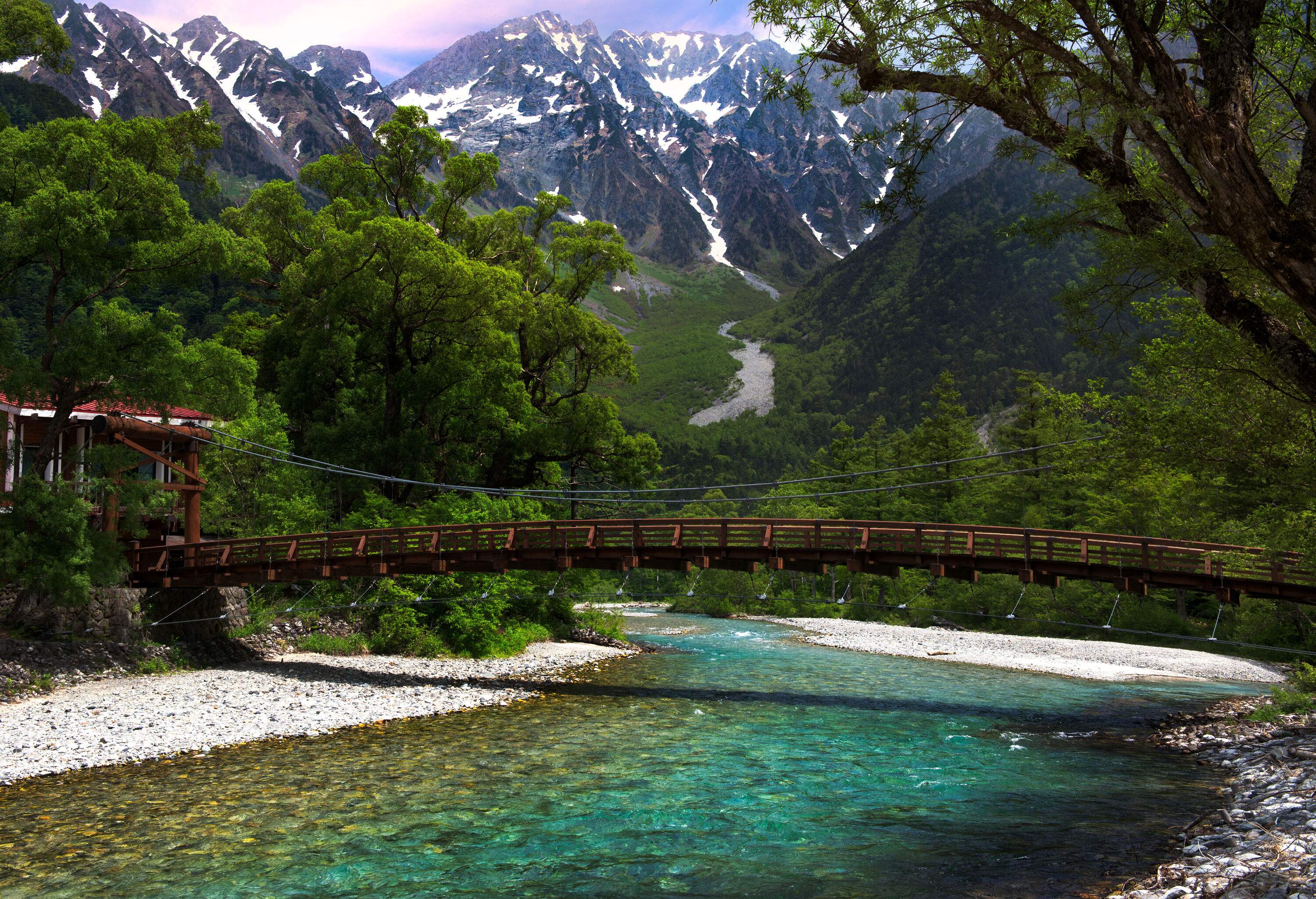 A semi-arch wooden suspension bridge over a crystal clear river with picturesque steep, sharp pointed mountains in the backdrop.
