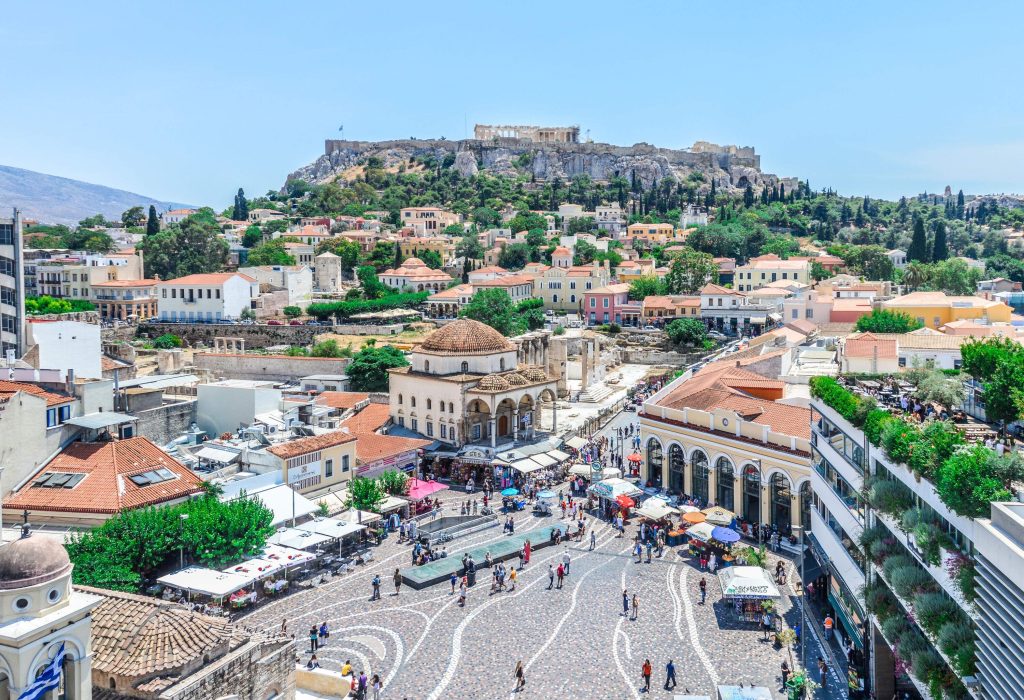 People strolling through a plaza surrounded by old town buildings with the Acropolis in the background.