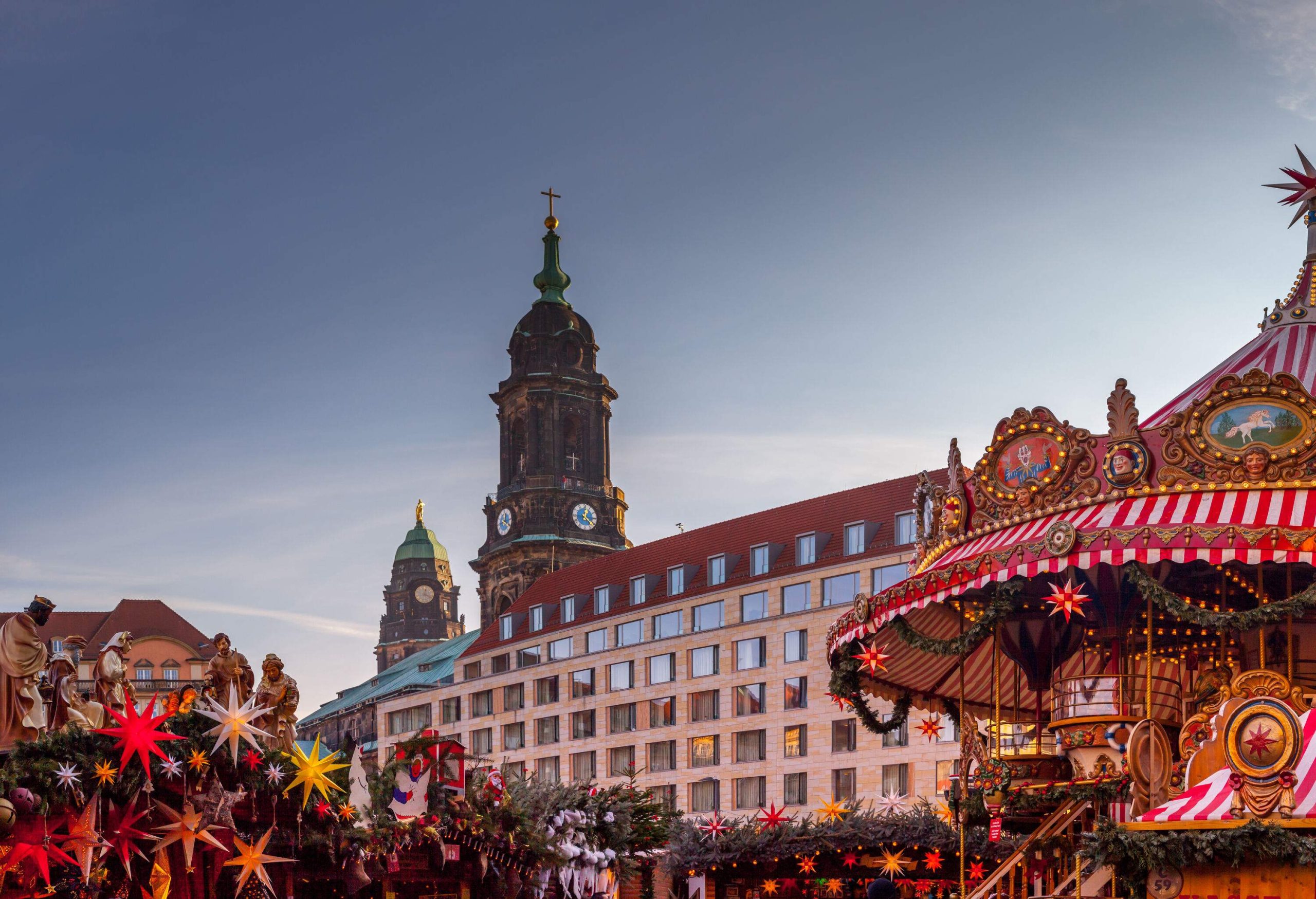 Shops decorated with a creche and lanterns beside a carousel in a Christmas market with a view of two dome towers in the background.