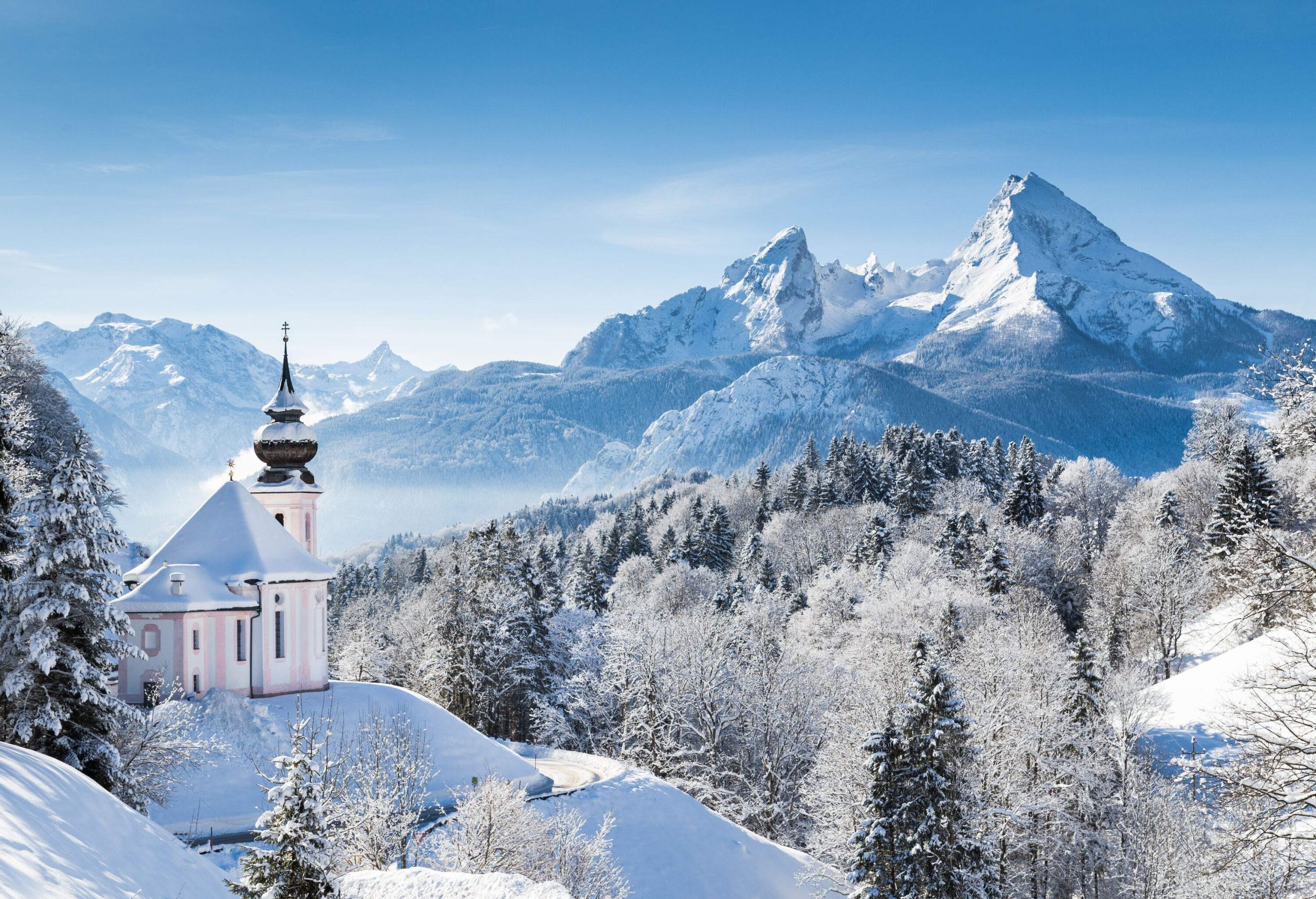 A small wayside church on an uphill road covered in snow and surrounded by trees and magnificent mountains.