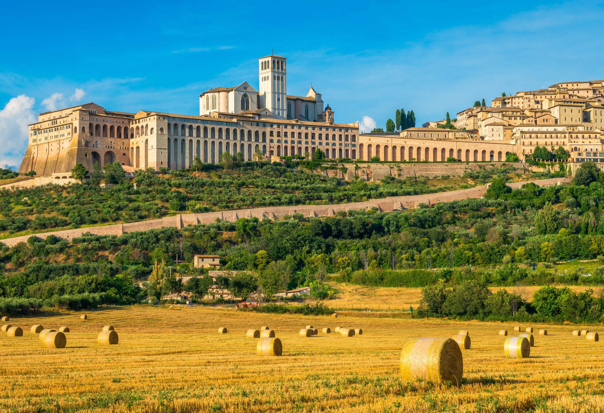 The majestic Basilica of Saint Francis and the town of Assisi overlooking a field strewn with hay bales.