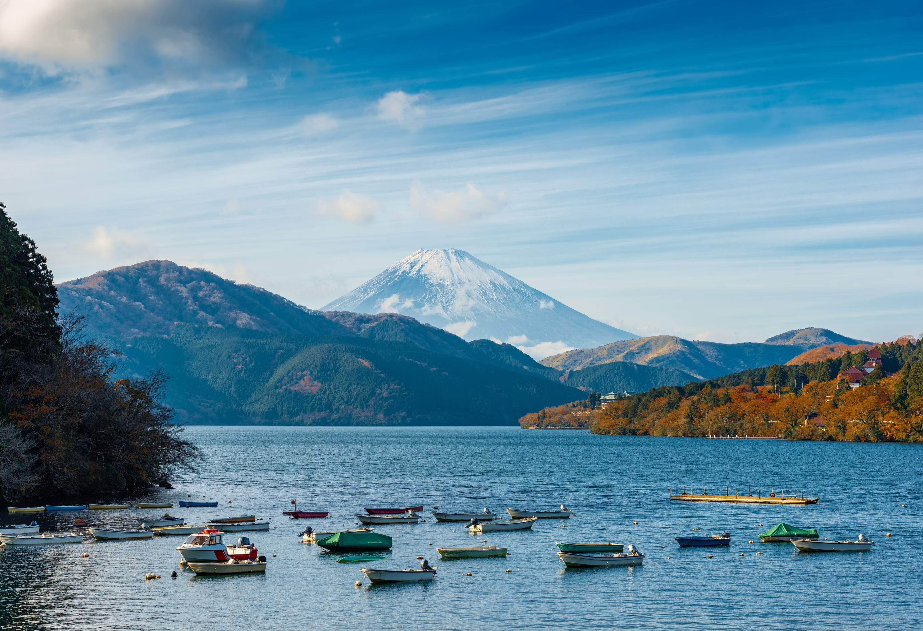 Several unoccupied boats on a lake, with the view of autumn trees on mountains and the snow-capped Mt. Fuji in the background.