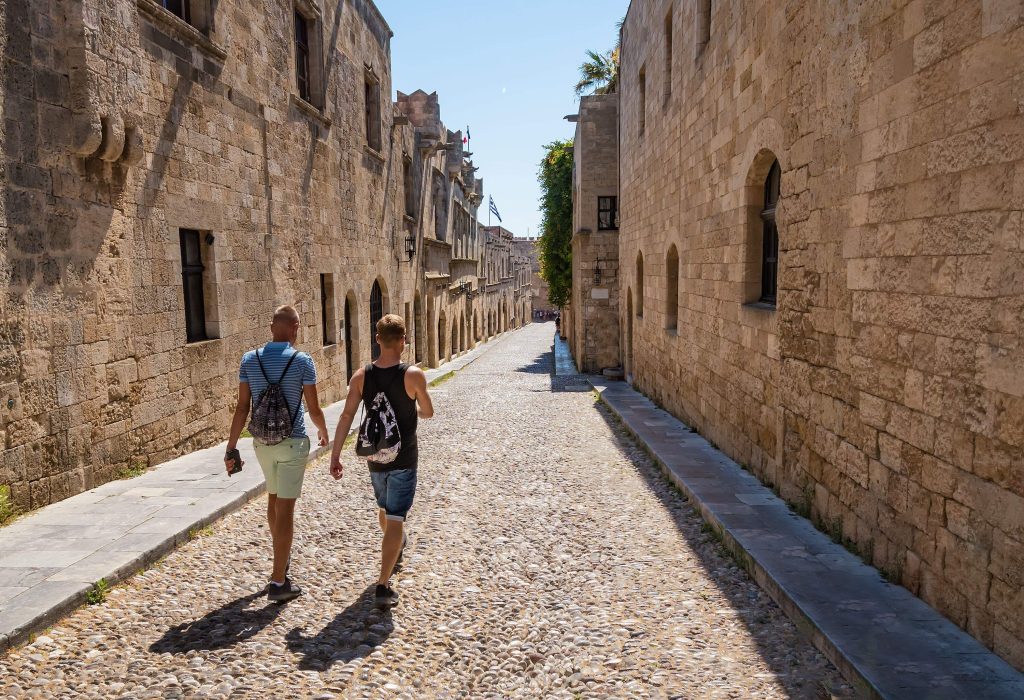 Two people walking down a stone-paved street lined with medieval structures.