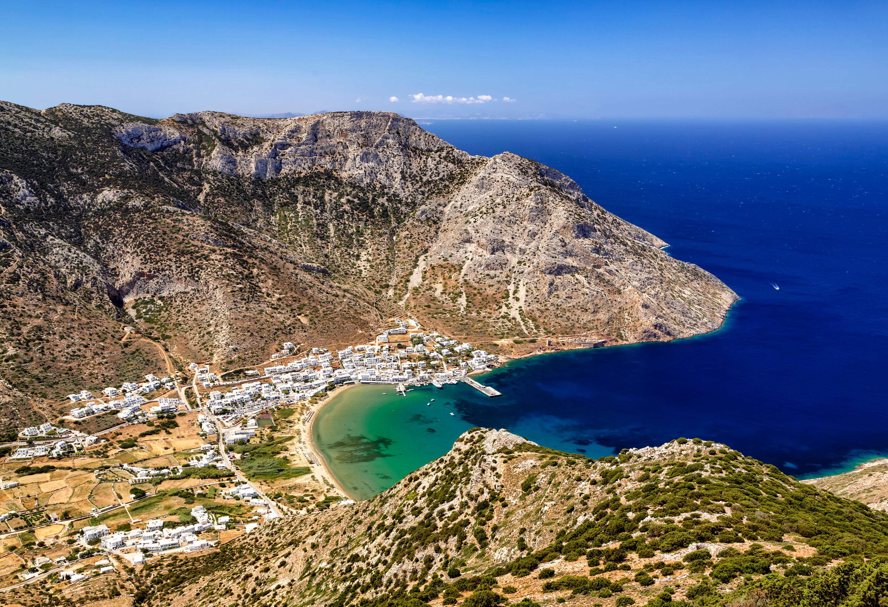 Aerial view of a white village and harbour among rocky hills with turquoise sea waters.