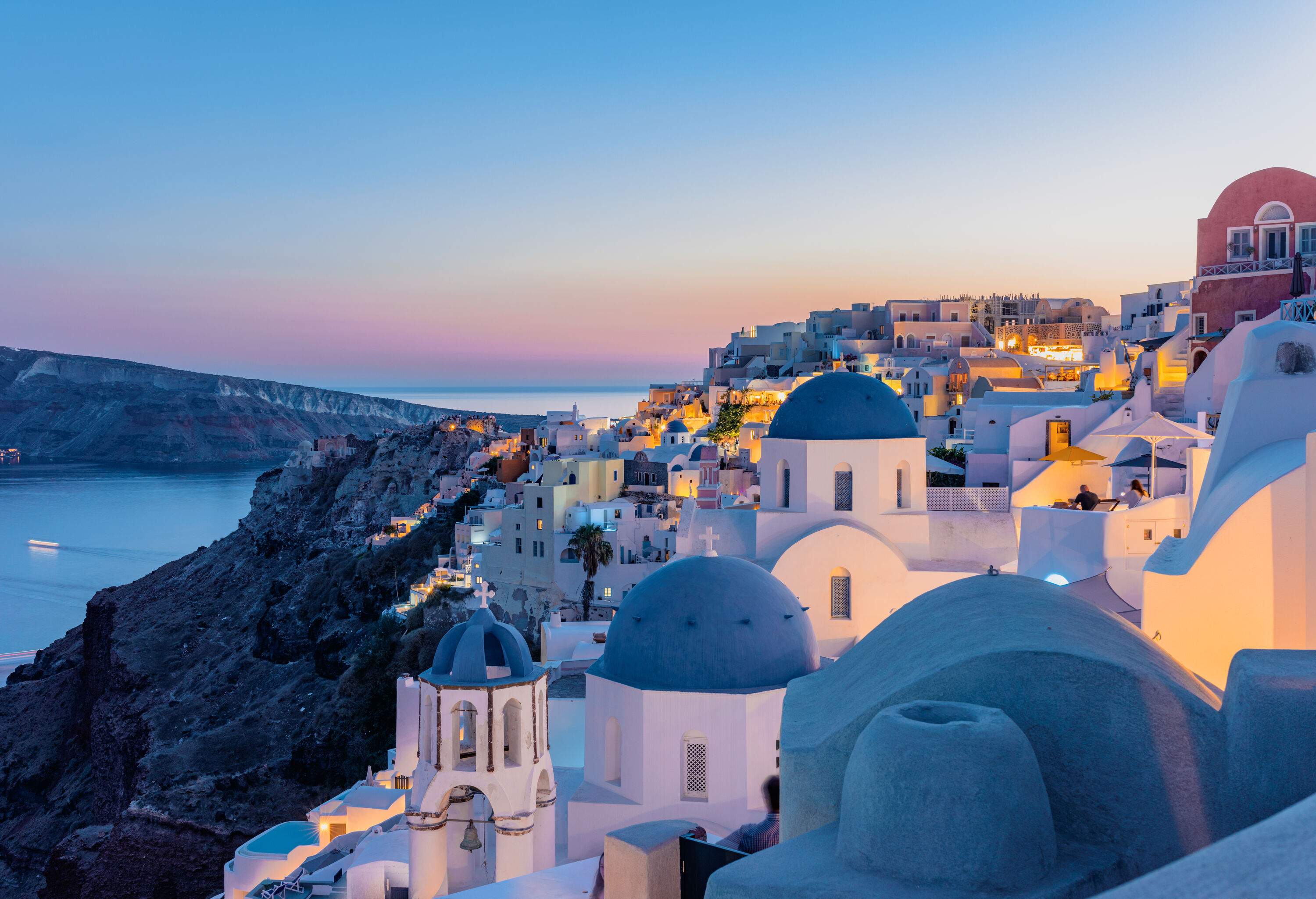 The soft blue hues of the sun setting over the sea cast a warm glow over the white buildings and blue-domed churches of the island's iconic architecture.