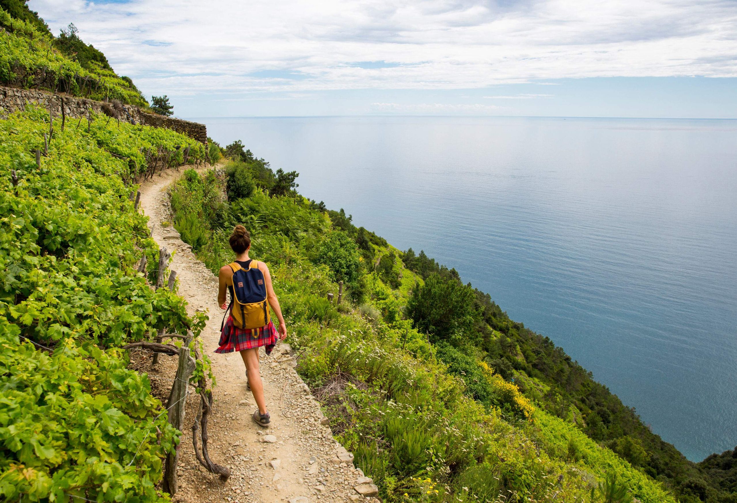 A person with a yellow backpack walking on a narrow sandy road of a mountain covered in lush greenery above the blue sea.
