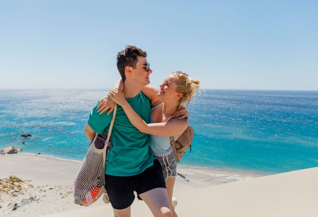 A young couple stands atop sand dunes at the beach, gazing lovingly into each other's eyes, with the man sporting a net shoulder bag and the woman wearing a spaghetti-strap blouse, creating a romantic and beachy atmosphere.