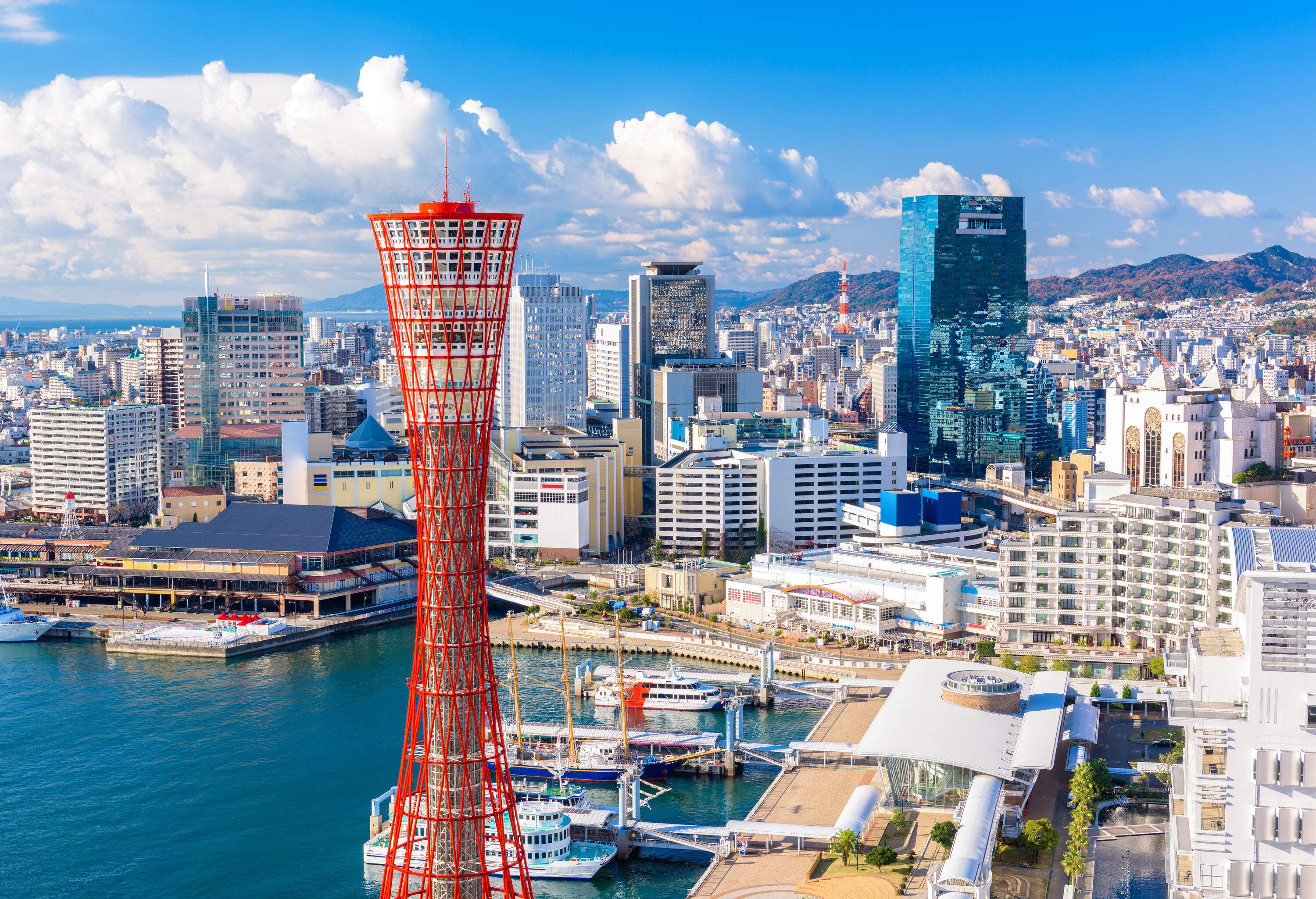 A red steel-truss tower overlooks a port with anchored boats and a cluster of buildings in a coastal city.