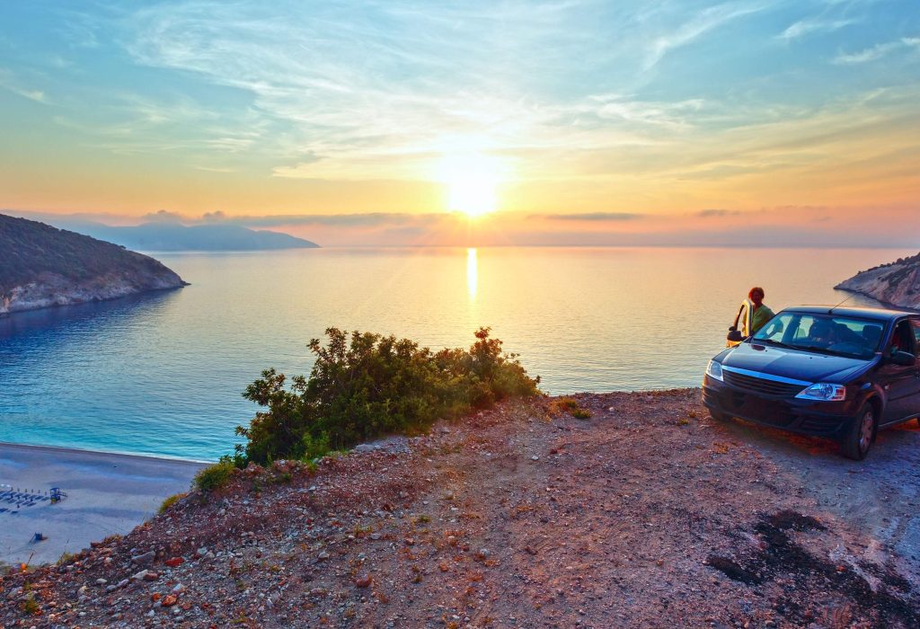 A woman standing behind an open door of a car parked on the beach with the beautiful sunset over the sea.