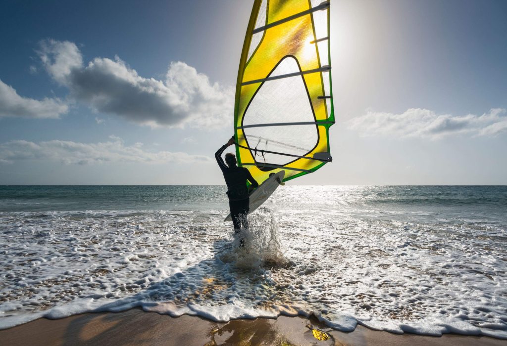 A person carrying windboarding equipment while heading towards the sea.