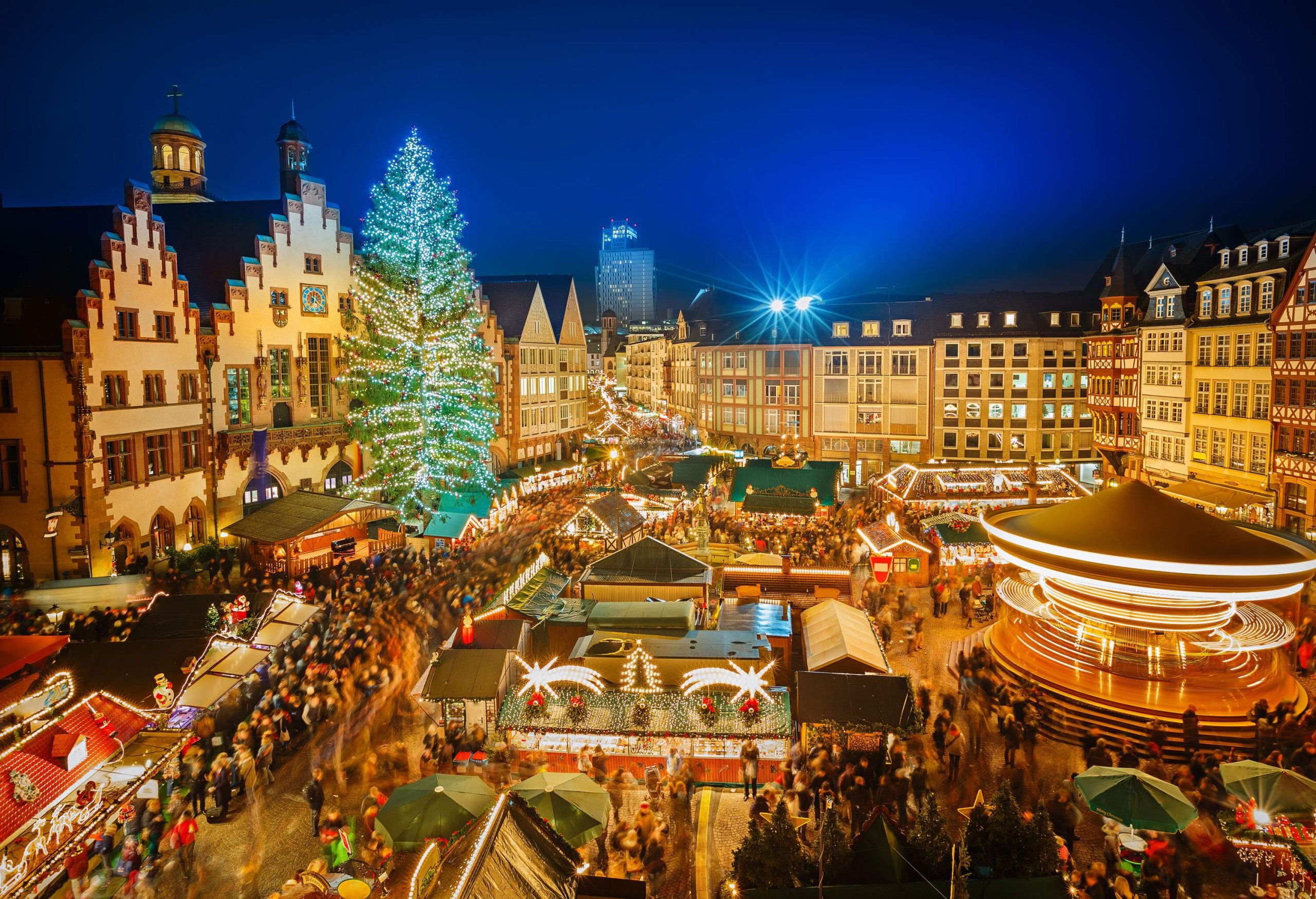 A brightly lit Christmas market packed with people and surrounded by eclectic structures.