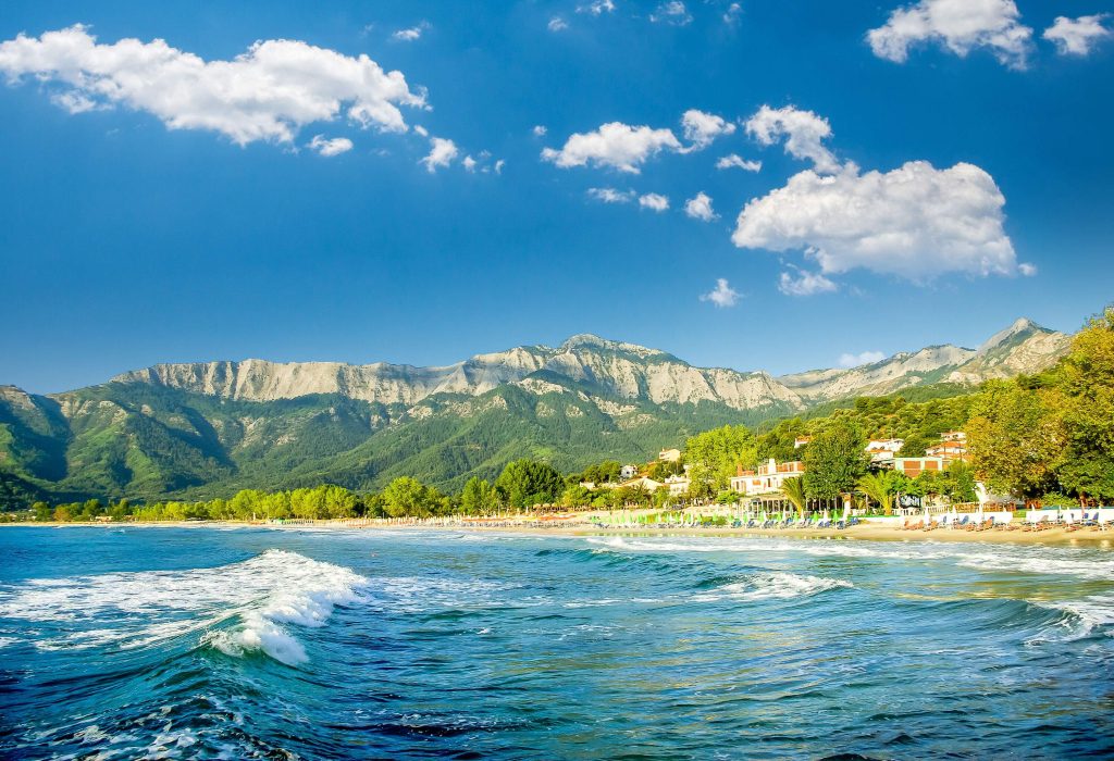 Waves rushing to the beach with trees and a stunning mountain range in the background.