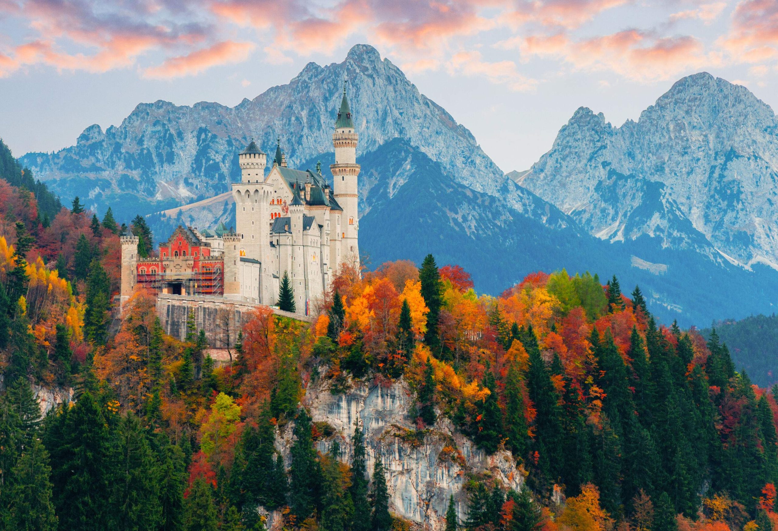 A medieval white castle with blue turrets on a rugged hill surrounded by trees with vivid autumn colours and a snow-capped mountain range in the background.