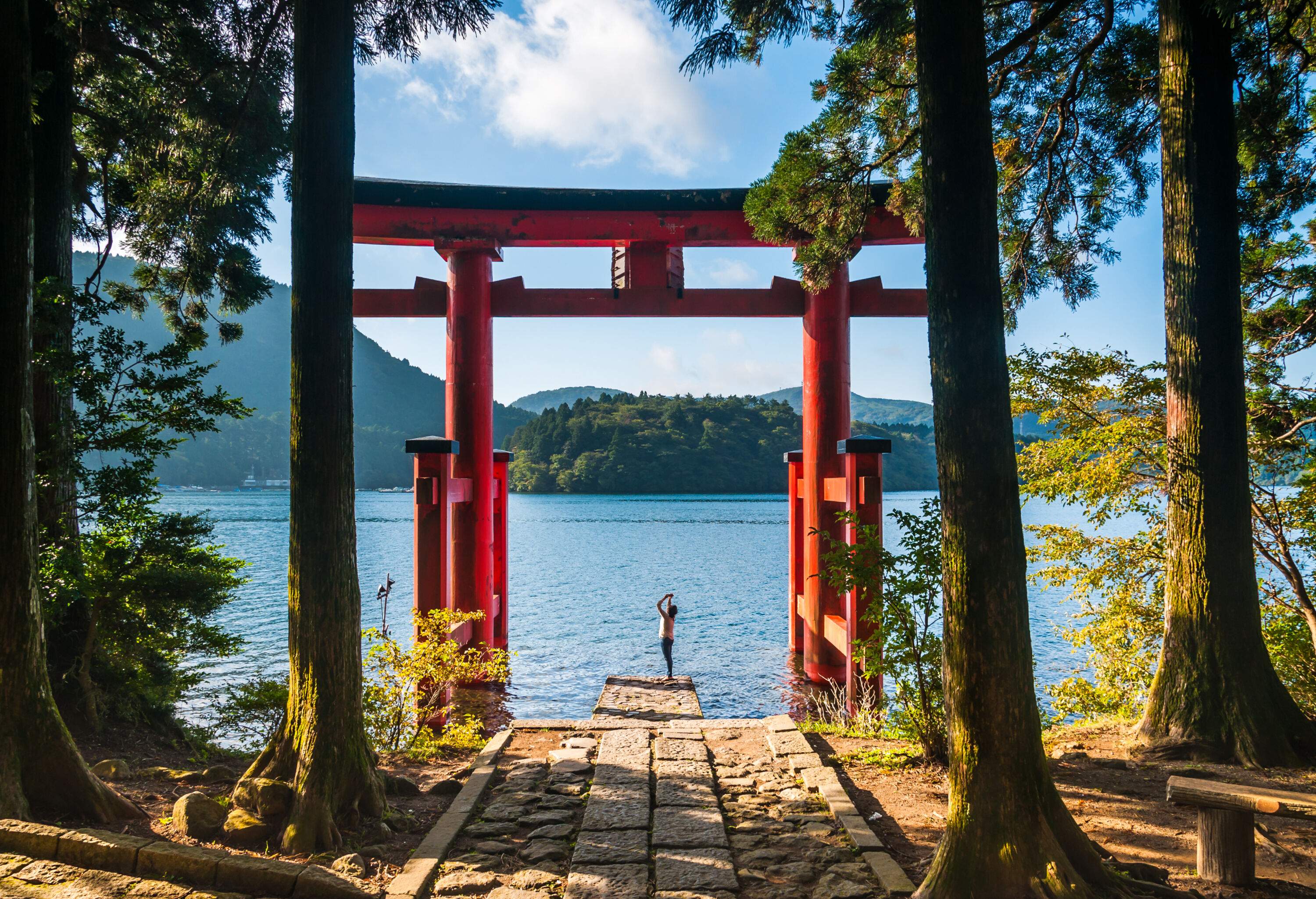 A young woman captures the beauty of a torii gate nestled among majestic trees against the backdrop of a tranquil lake through her phone's lens.