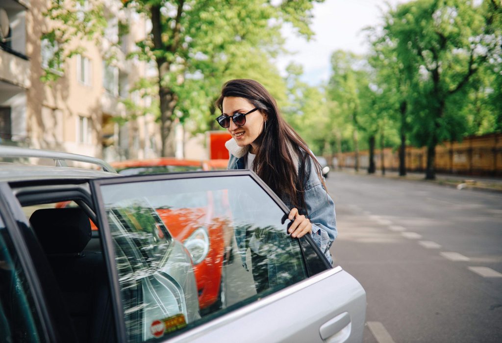 A woman wearing sunglasses smiles as she opens a car's door.