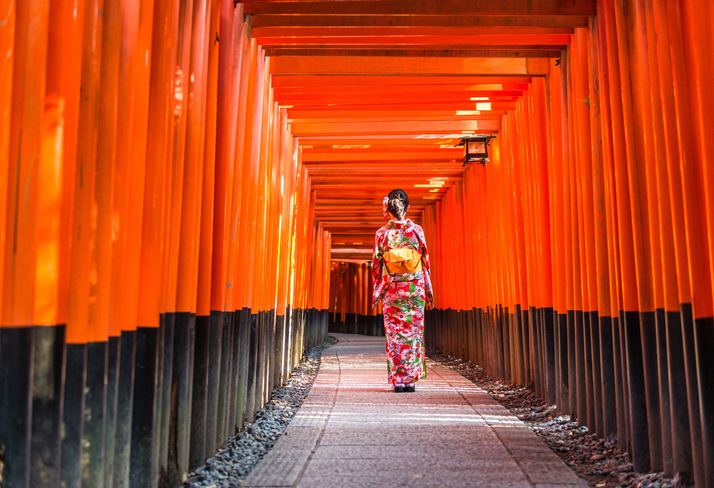 A woman in a traditional and colourful kimono on a pathway lined with orange pillars with Japanese inscriptions.