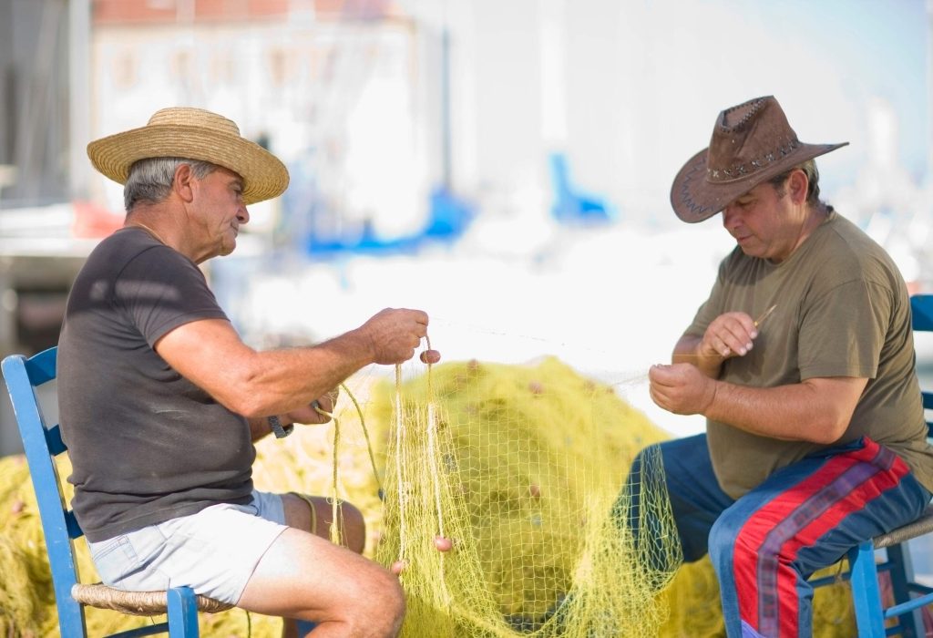 Two men in hats sit on blue wooden chairs and mend yellow fishing nets.