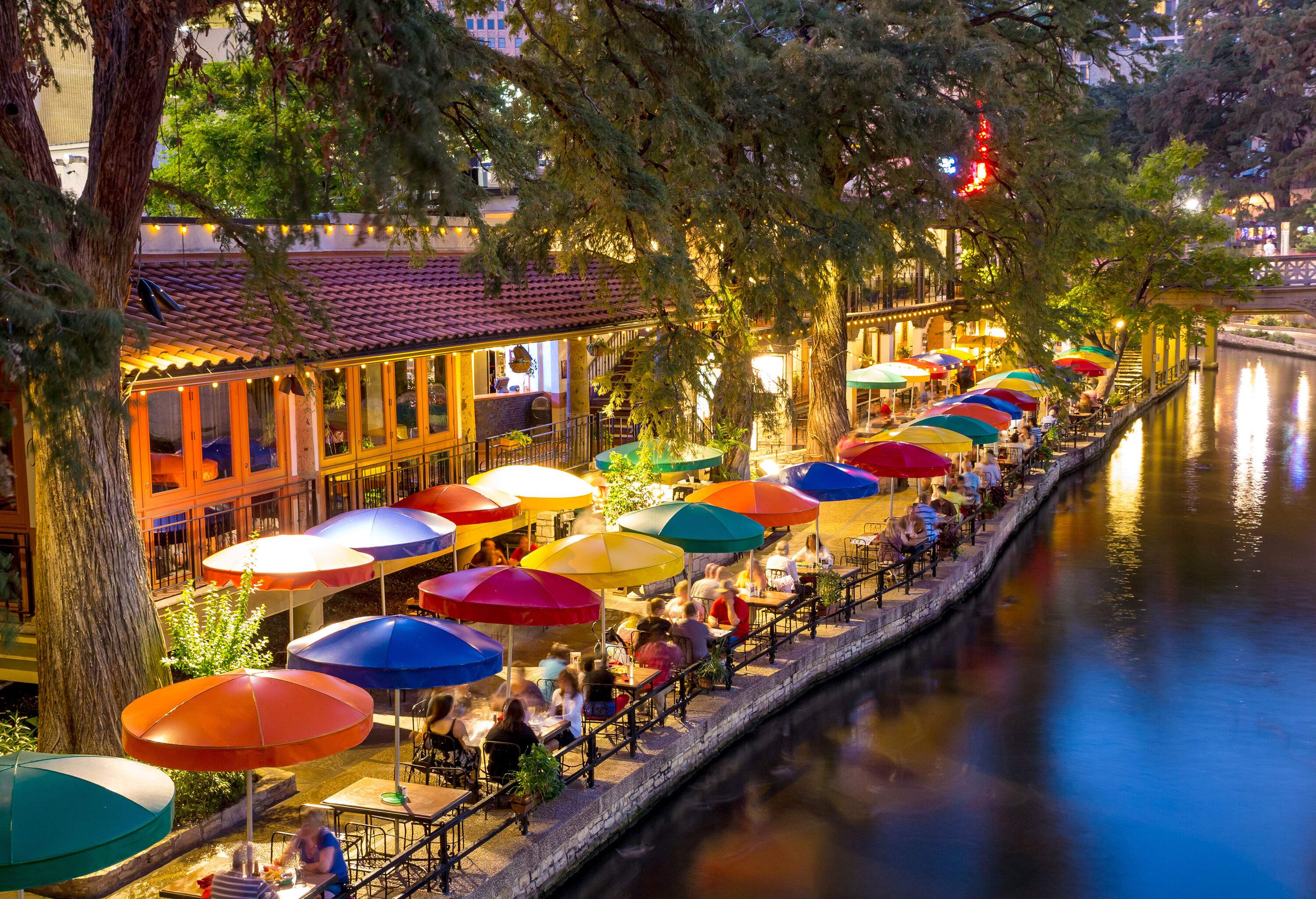 A scenic riverwalk unfolds, featuring rows of vibrant parasols and buildings with inviting outdoor dining, all set amidst a backdrop of towering trees.