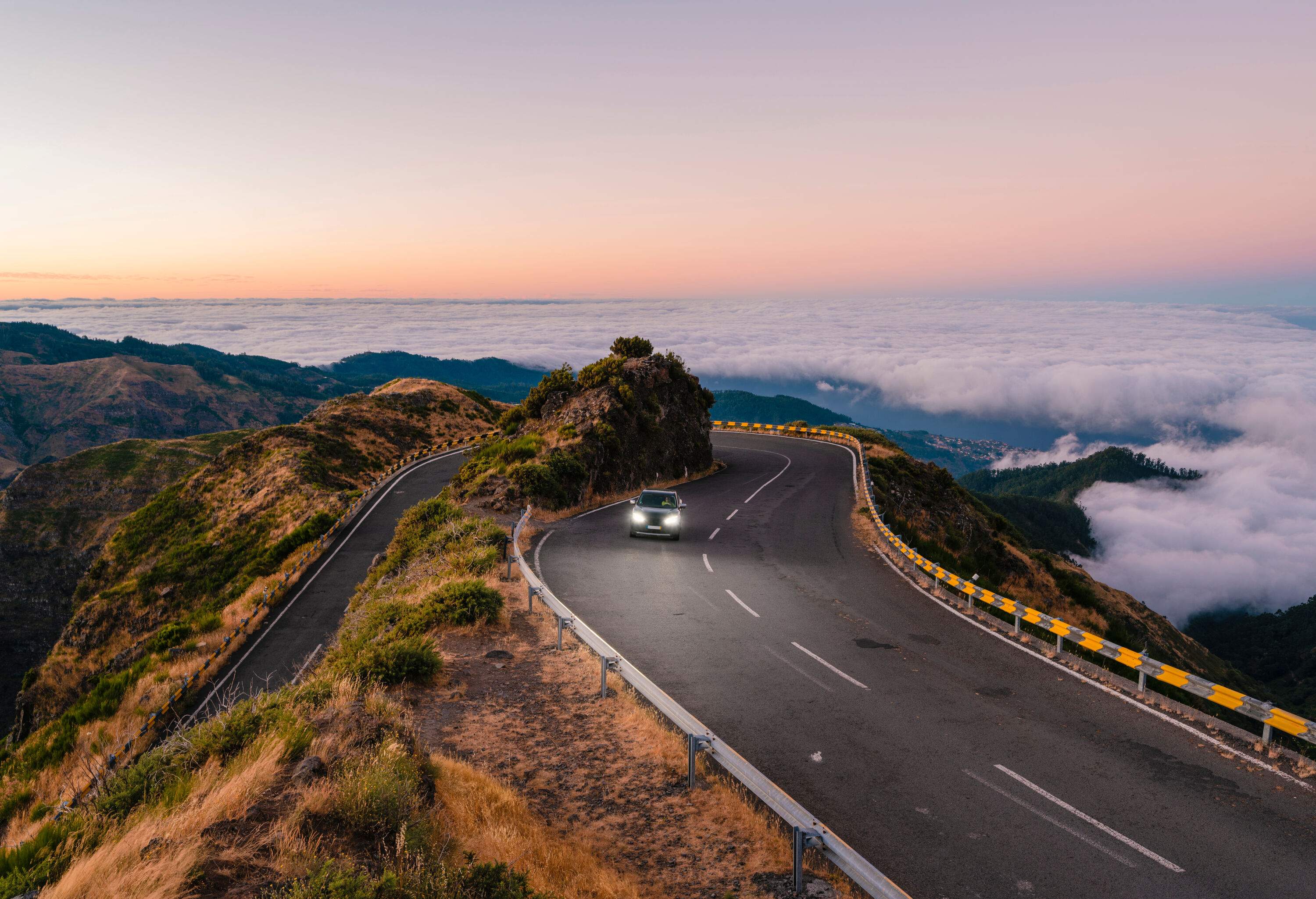 A car on a winding mountain road with a background of a sea of clouds covering the mountain range.