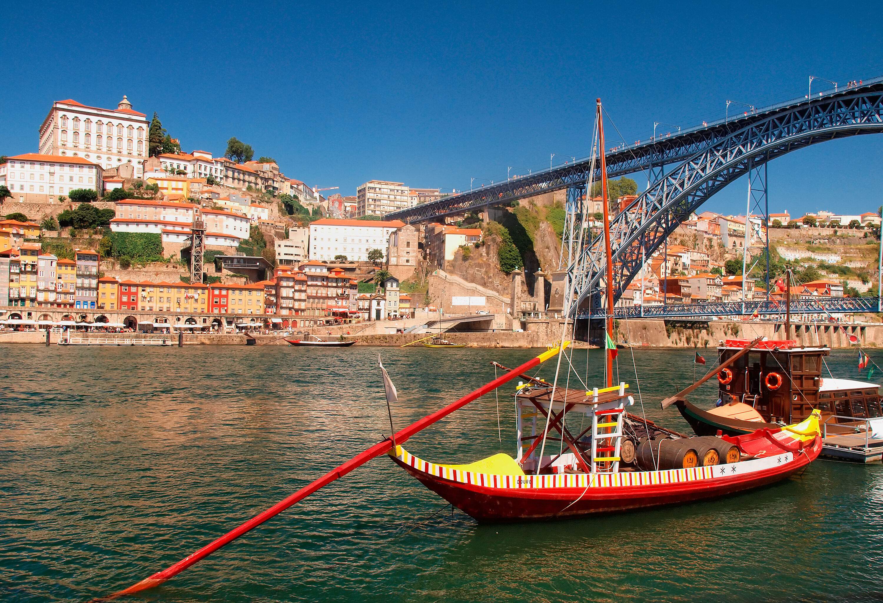 A rabelo boat about to pass the double-deck metal arch bridge across the river linked to a hill of tall buildings.
