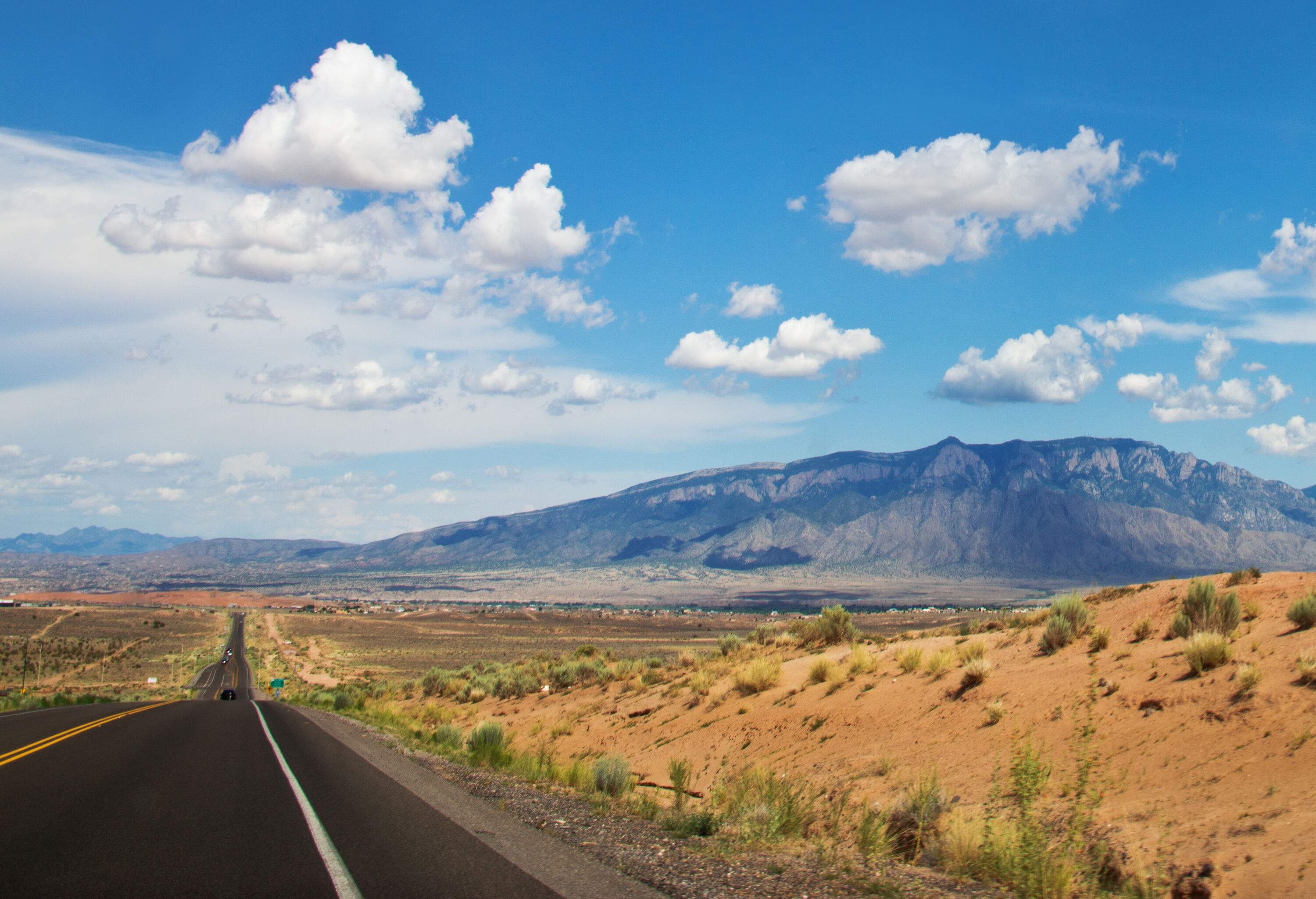 A long paved highway on a barren landscape overlooking the mountain range against the cloudy blue sky.