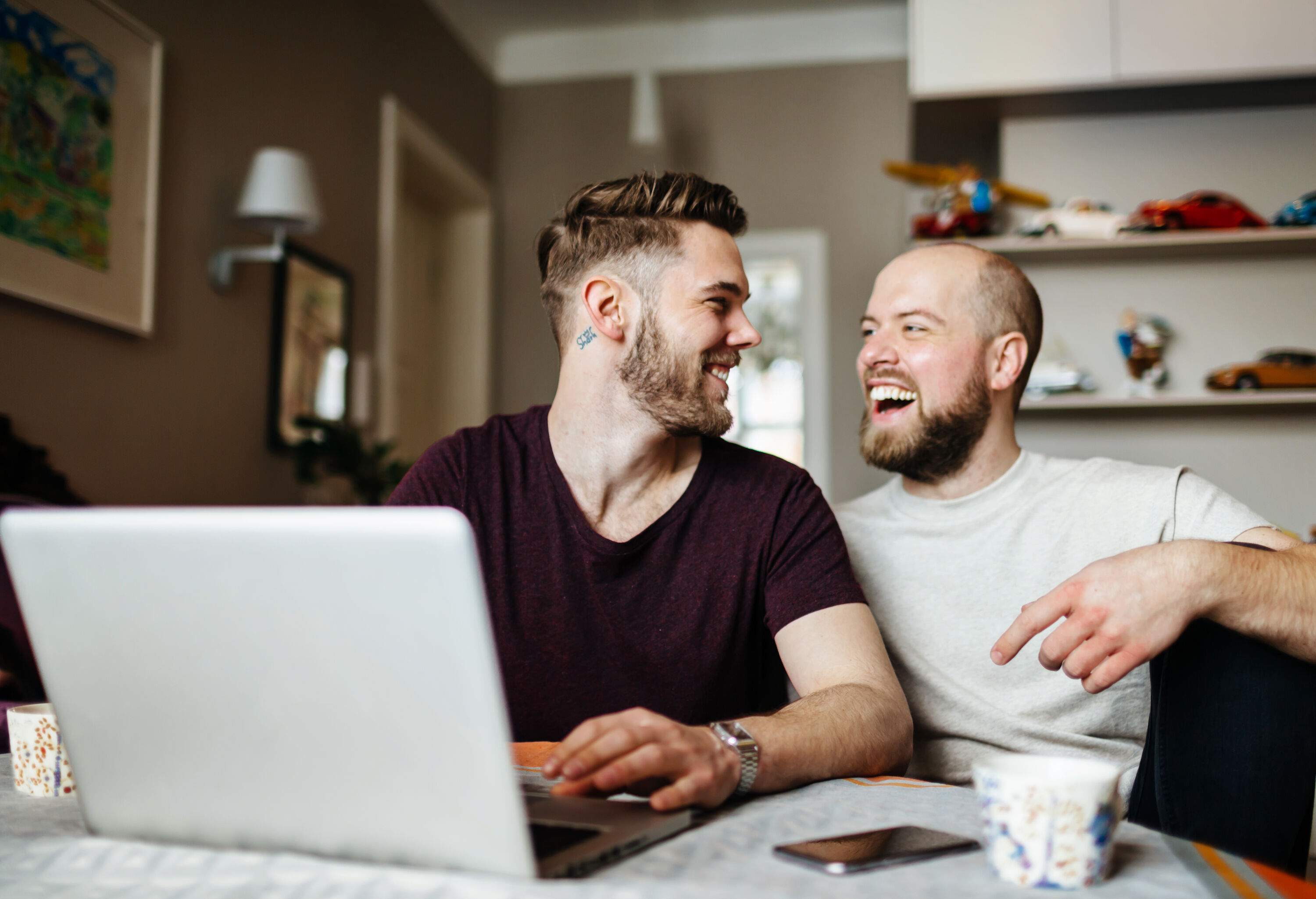 A happy gay couple is sitting in the dining area and holding a laptop in front of them.