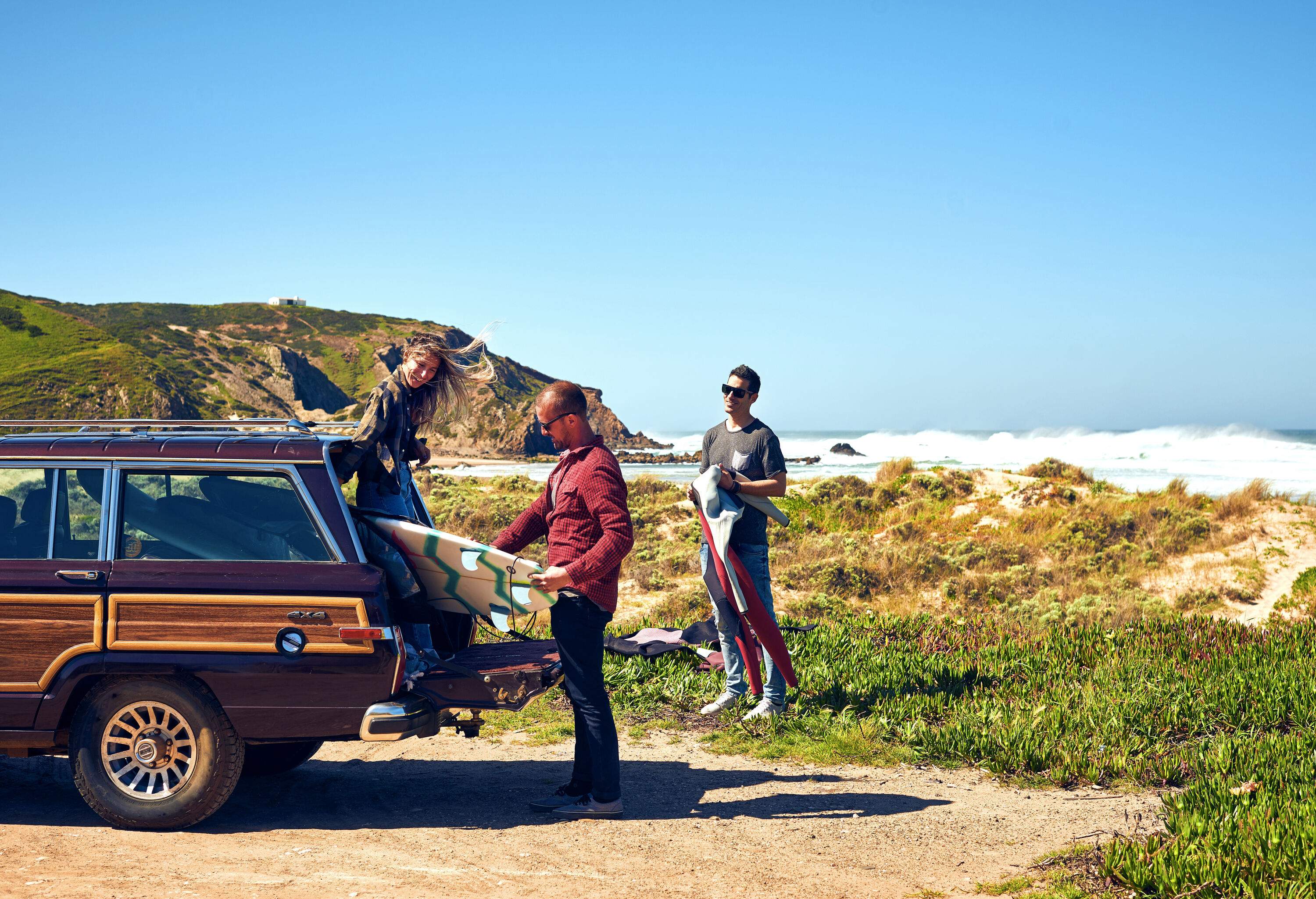 Three friends unloading their surfboards from a car's trunk in a grassy beach.