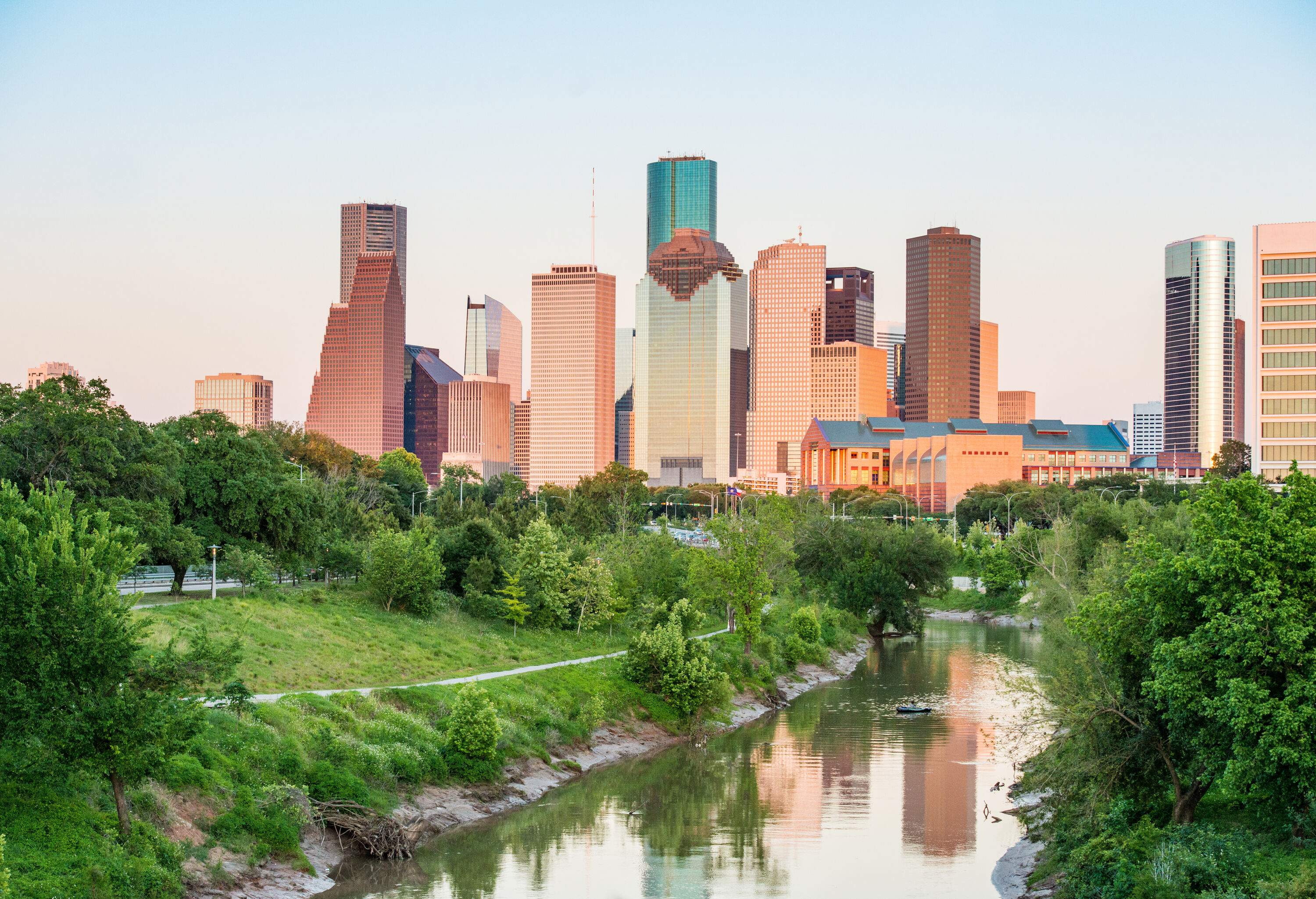 A stream guarded by lush green trees directed towards a cluster of compact tall buildings against the white sky.