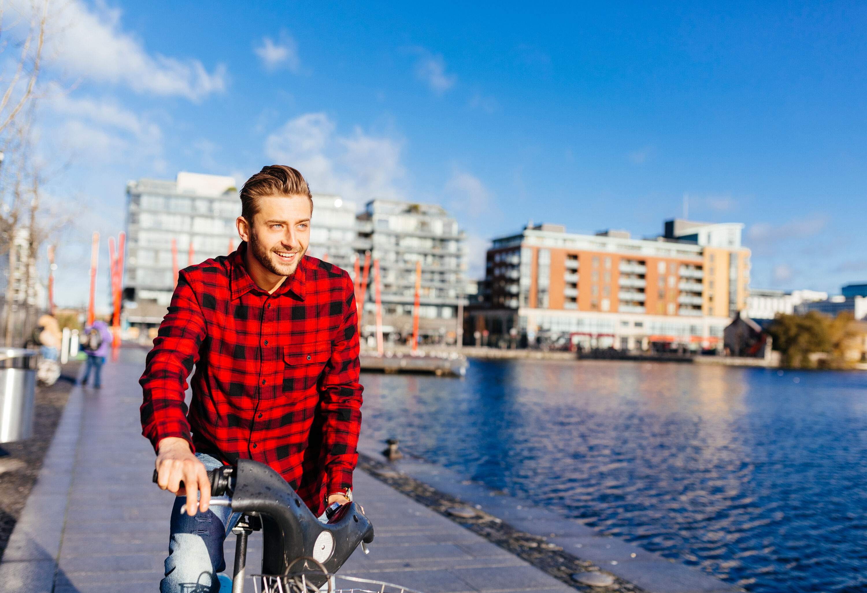 A young man in a red flannel shirt riding a bike along a canal in the city, with buildings in the background.