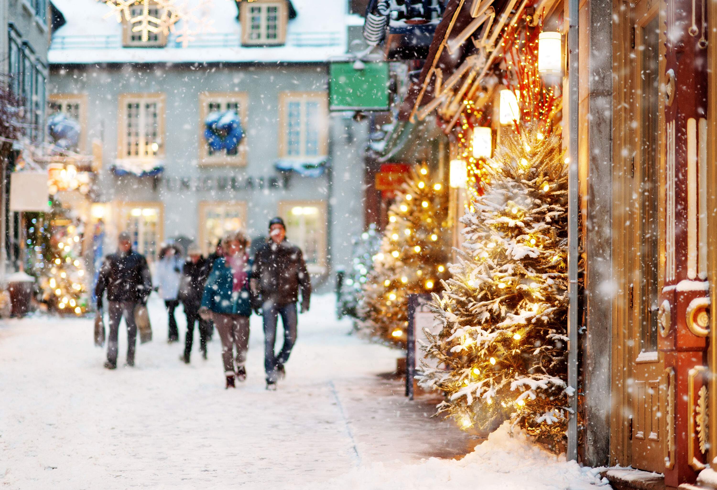A blurred image of people walking along the road covered with snow, alongside illuminated Christmas trees outside the stores on a cold winter night.