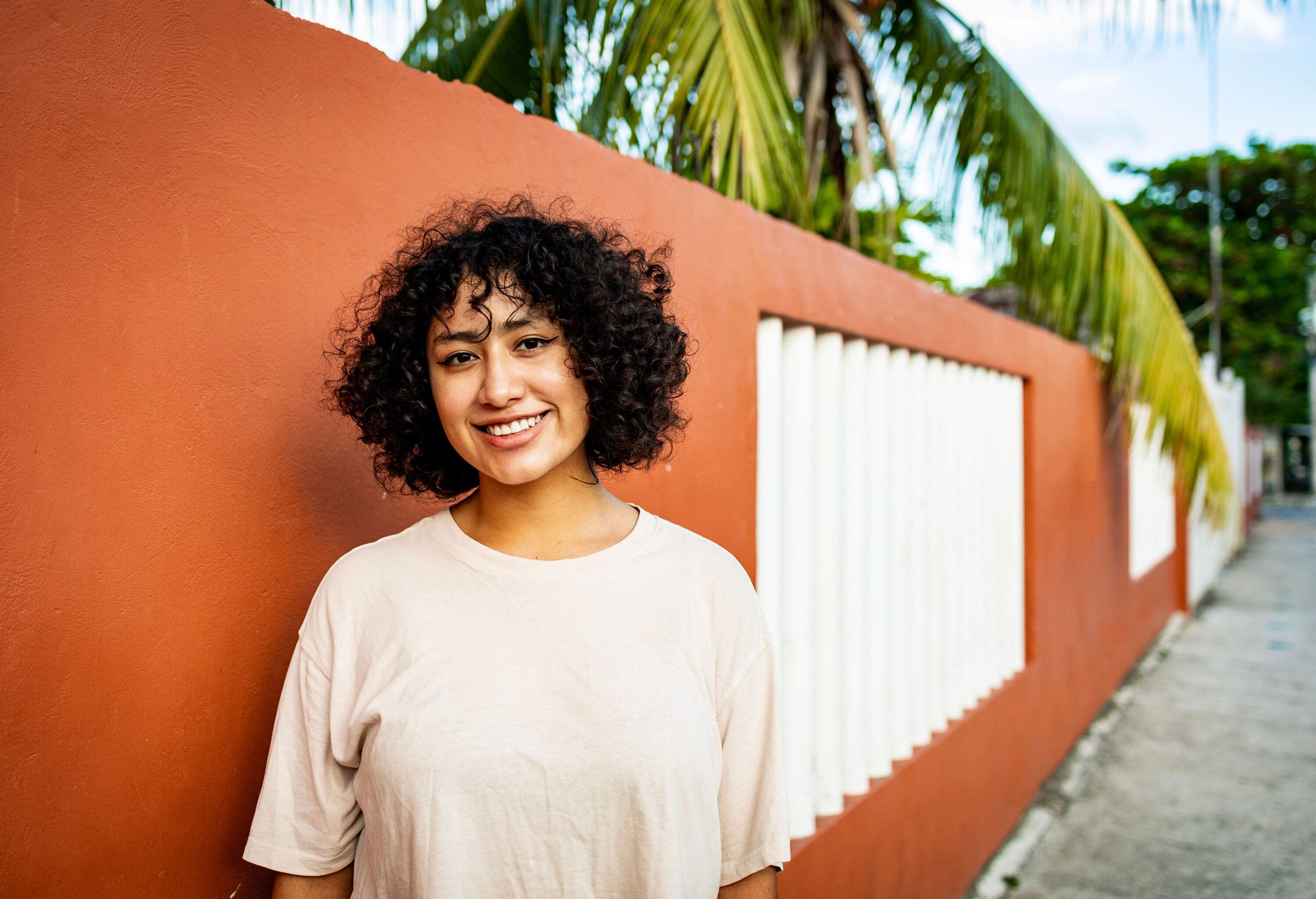 A young woman with curly hair smiling as she stands on the sidewalk next to an orange wall.