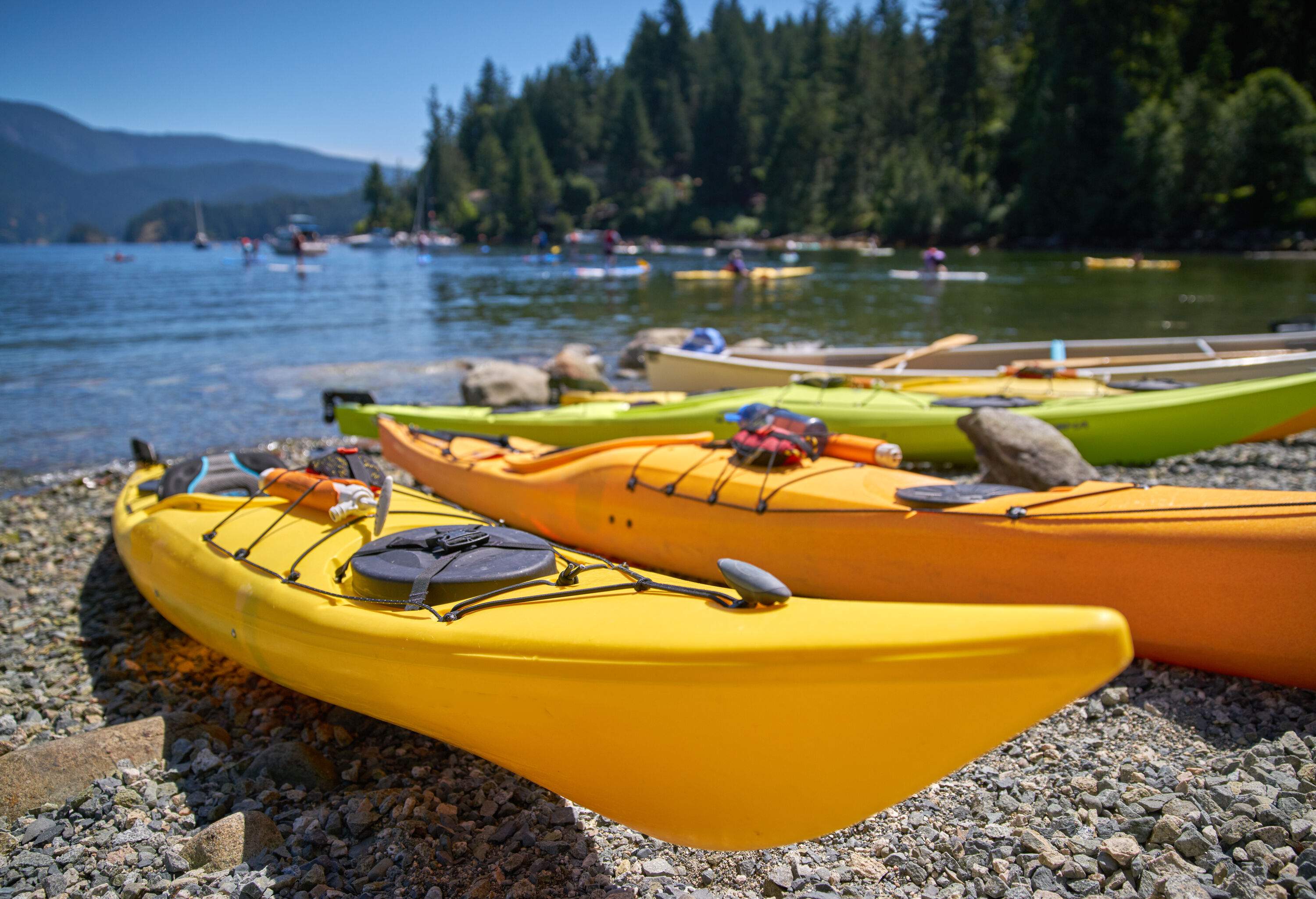 Yellow and green canoes docked on the pebbly shore of a bay, with people kayaking in the background.
