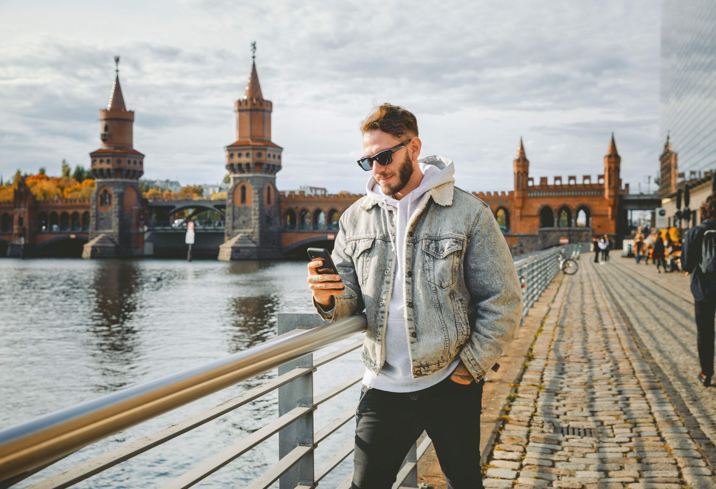 A man in sunglasses, denim jacket, and black pants leans on a promenade railing while using his smartphone.