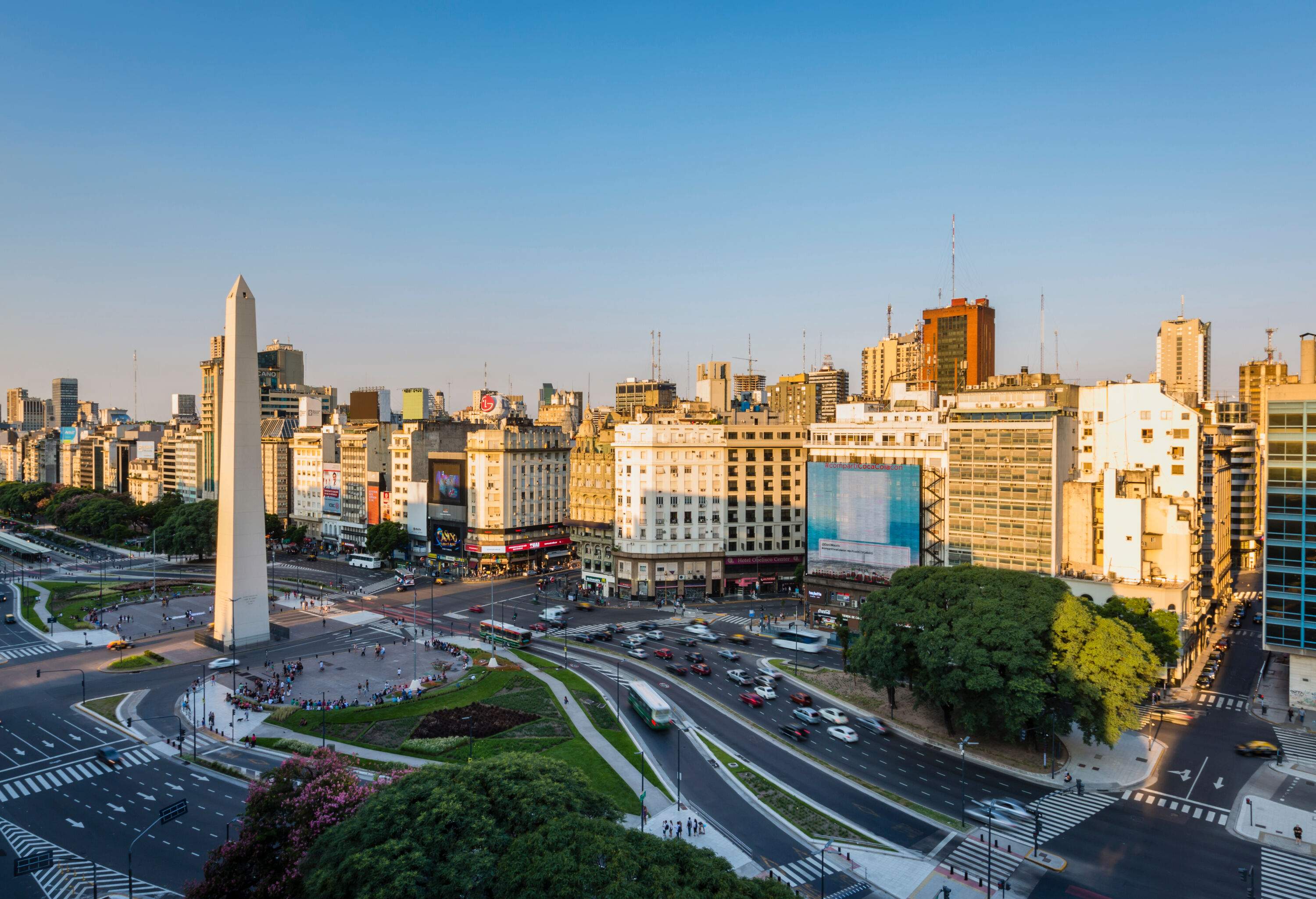 A white stone tower monument perched in the intersection of bustling avenues lined with compact tall buildings.