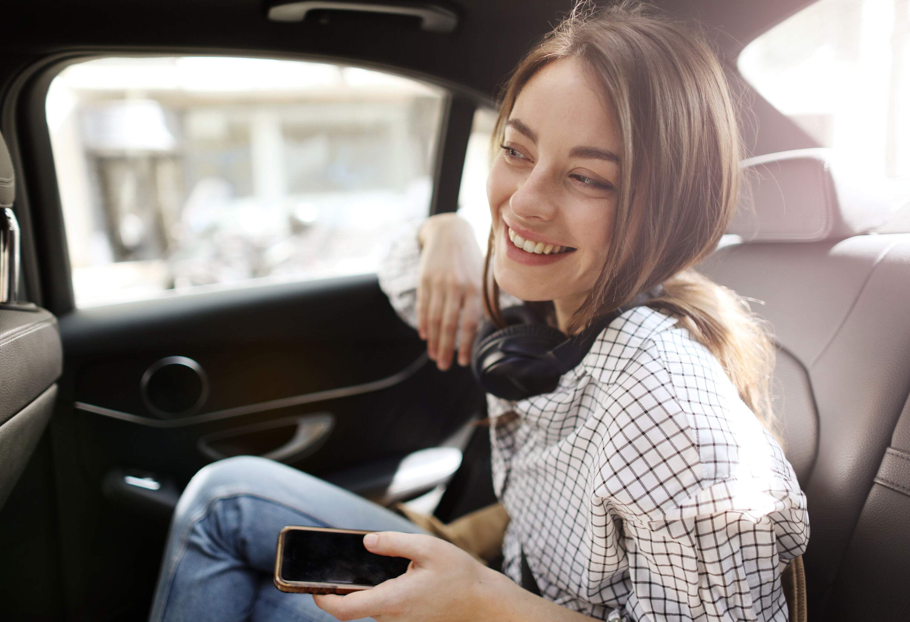 A brunette woman in a white chequered shirt smiles as she sits at the back of a car.