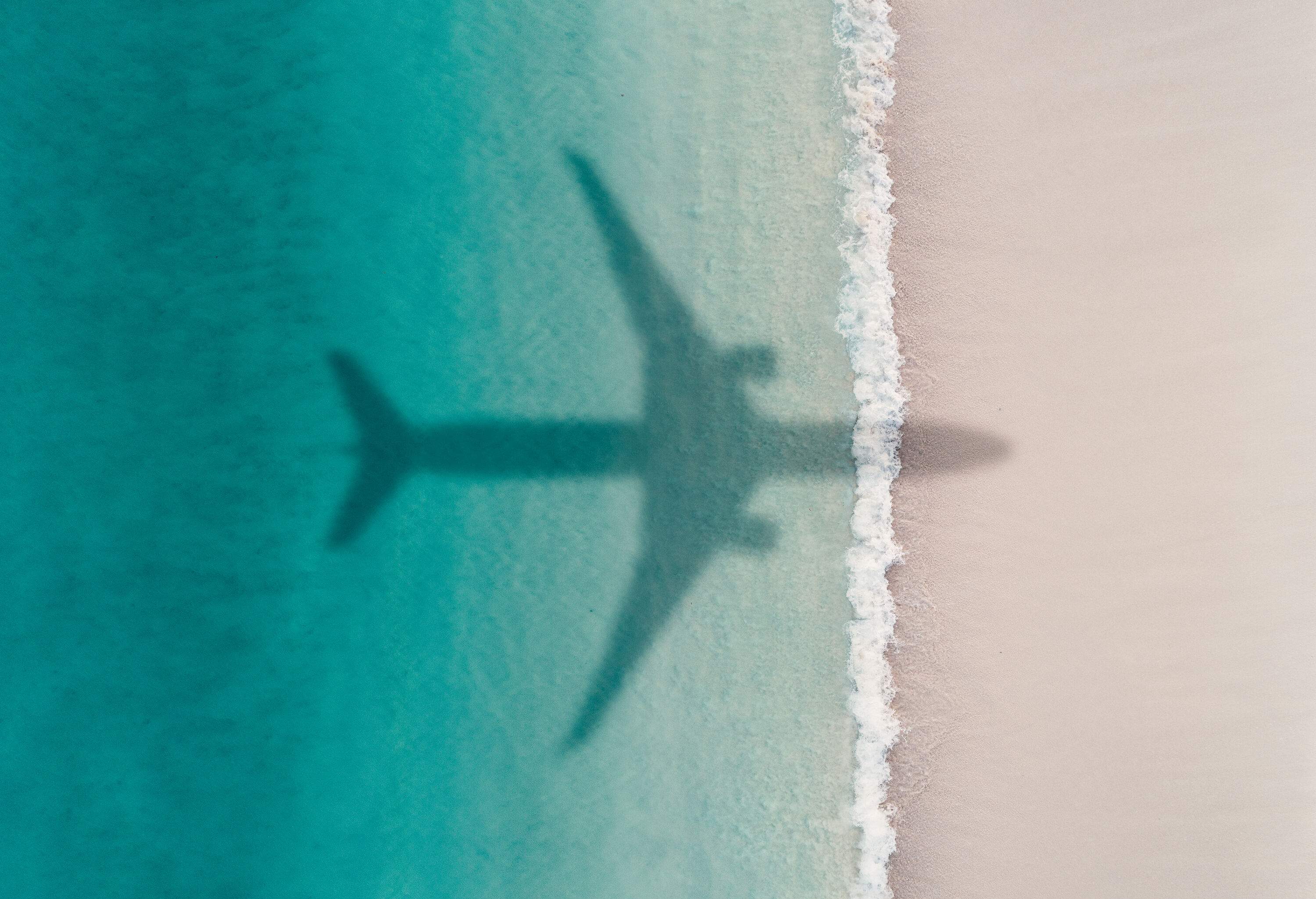 An airplane's shadow casts on a beach with turquoise waters.