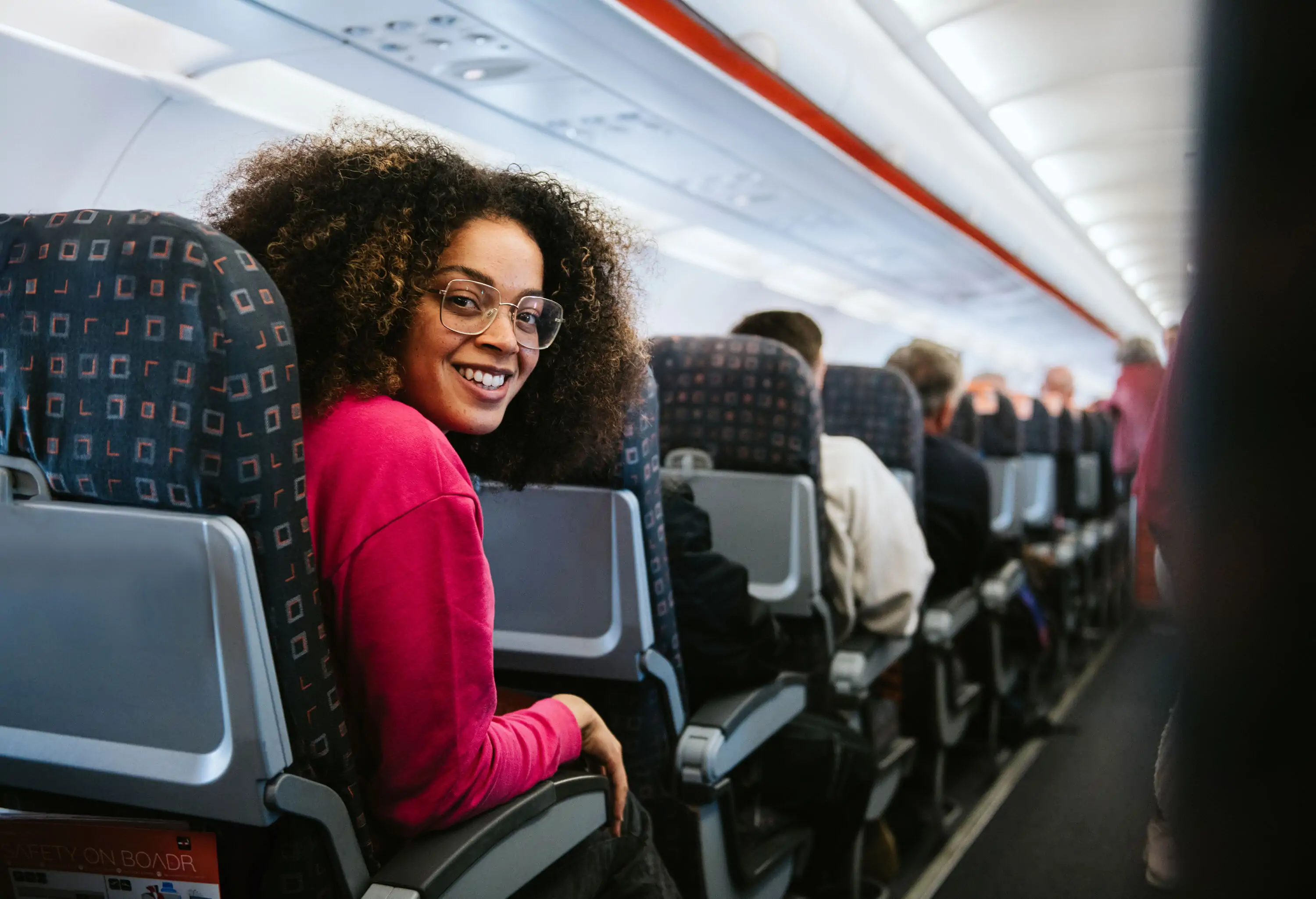 A happy young woman sits in a seat in a commercial airplane. She glances over her shoulder and smiles at the camera. She looks confident and excited to be on the flight.
