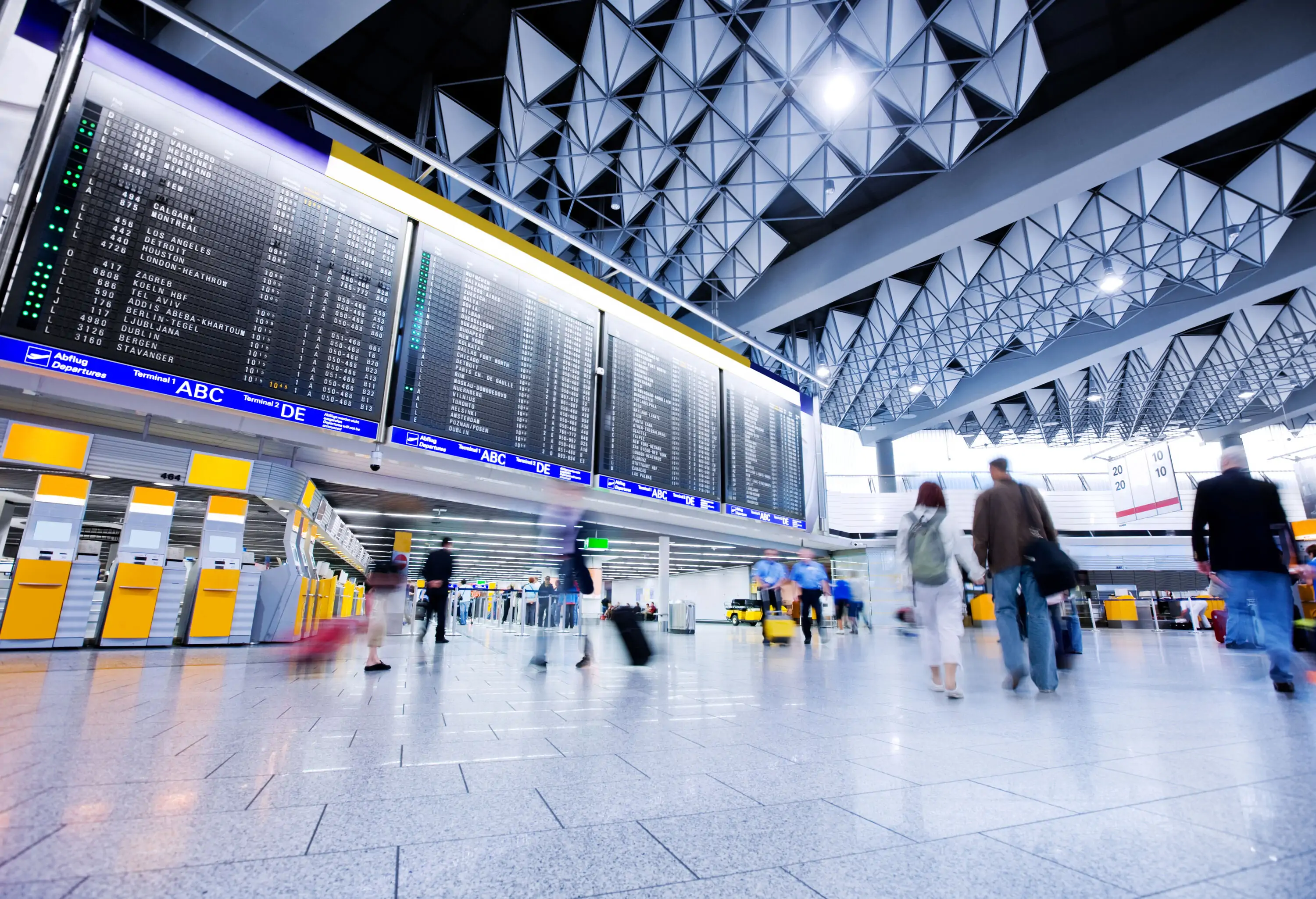A crowded airport hallway with flight information LED screens above the check-in counters.