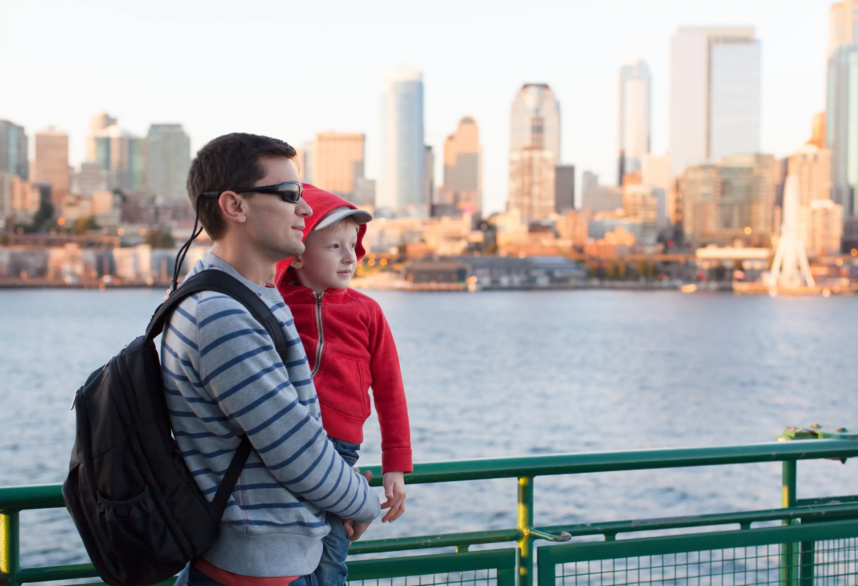 A man in a striped sweater carries a child in a red hoodie with views of the riverside urban scene.