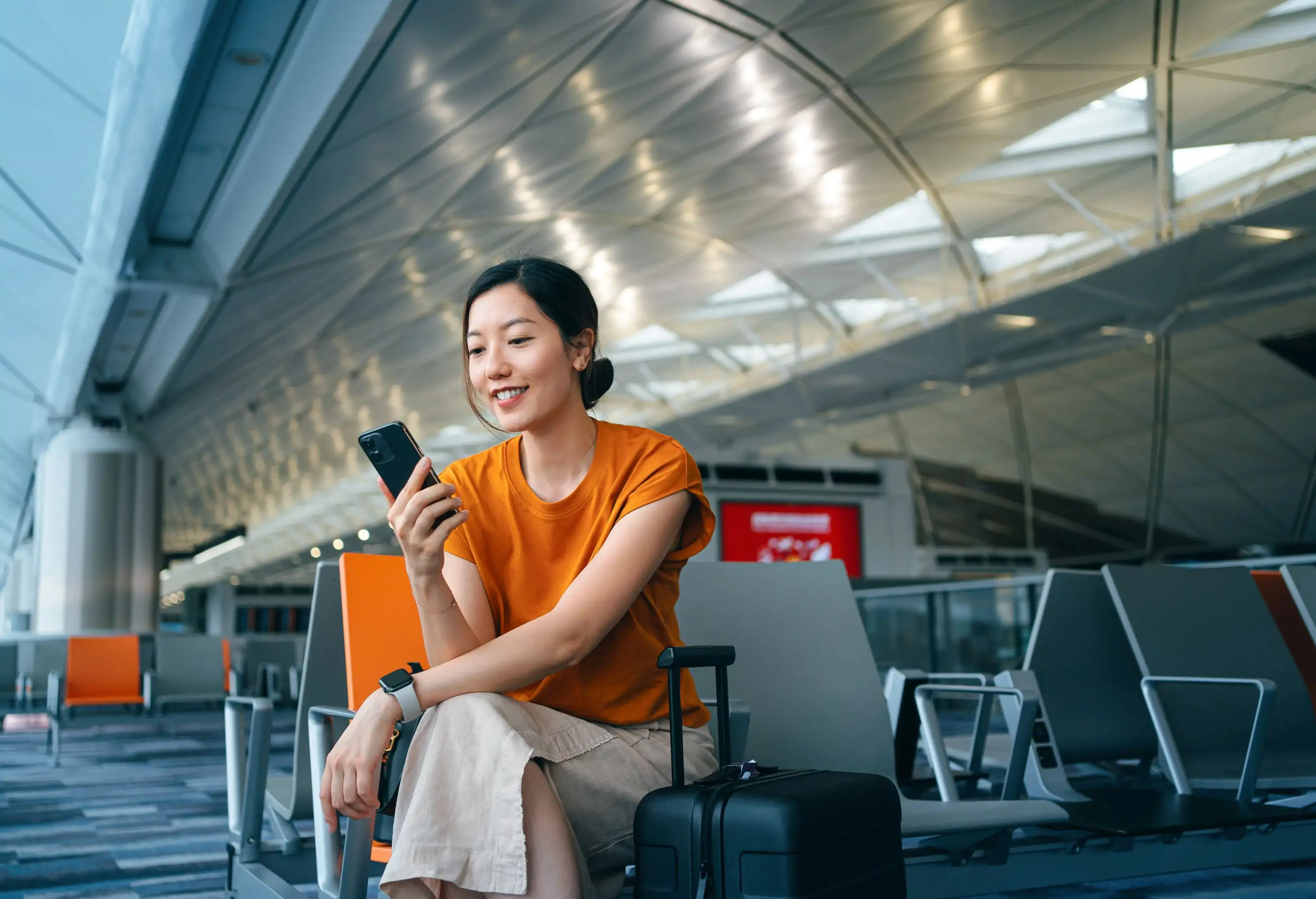Young Asian woman with suitcase using smartphone while waiting for her flight at airport terminal.
