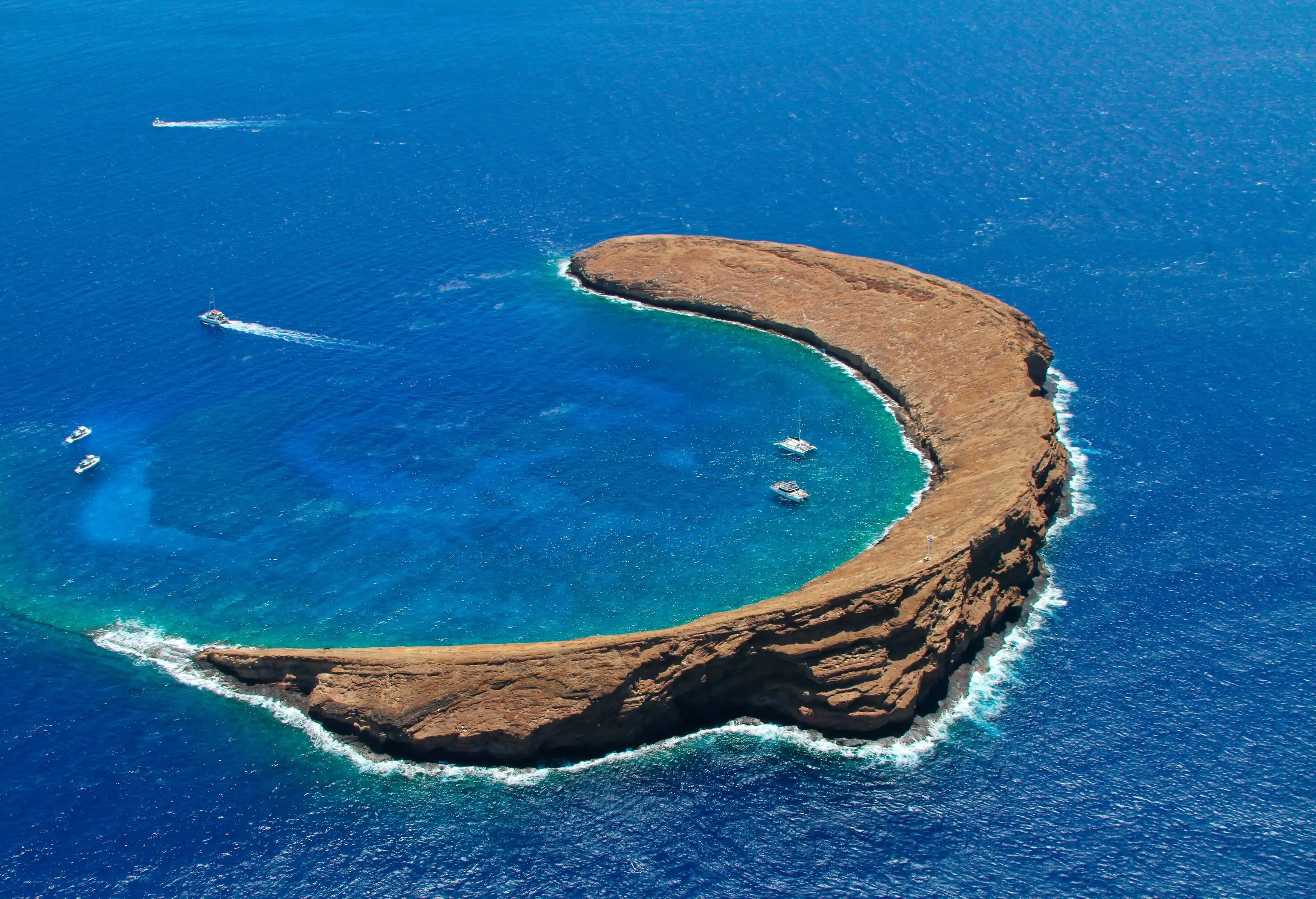 Aerial view of a half-moon crater swallowed by the ocean with boats and snorkelers in its bay