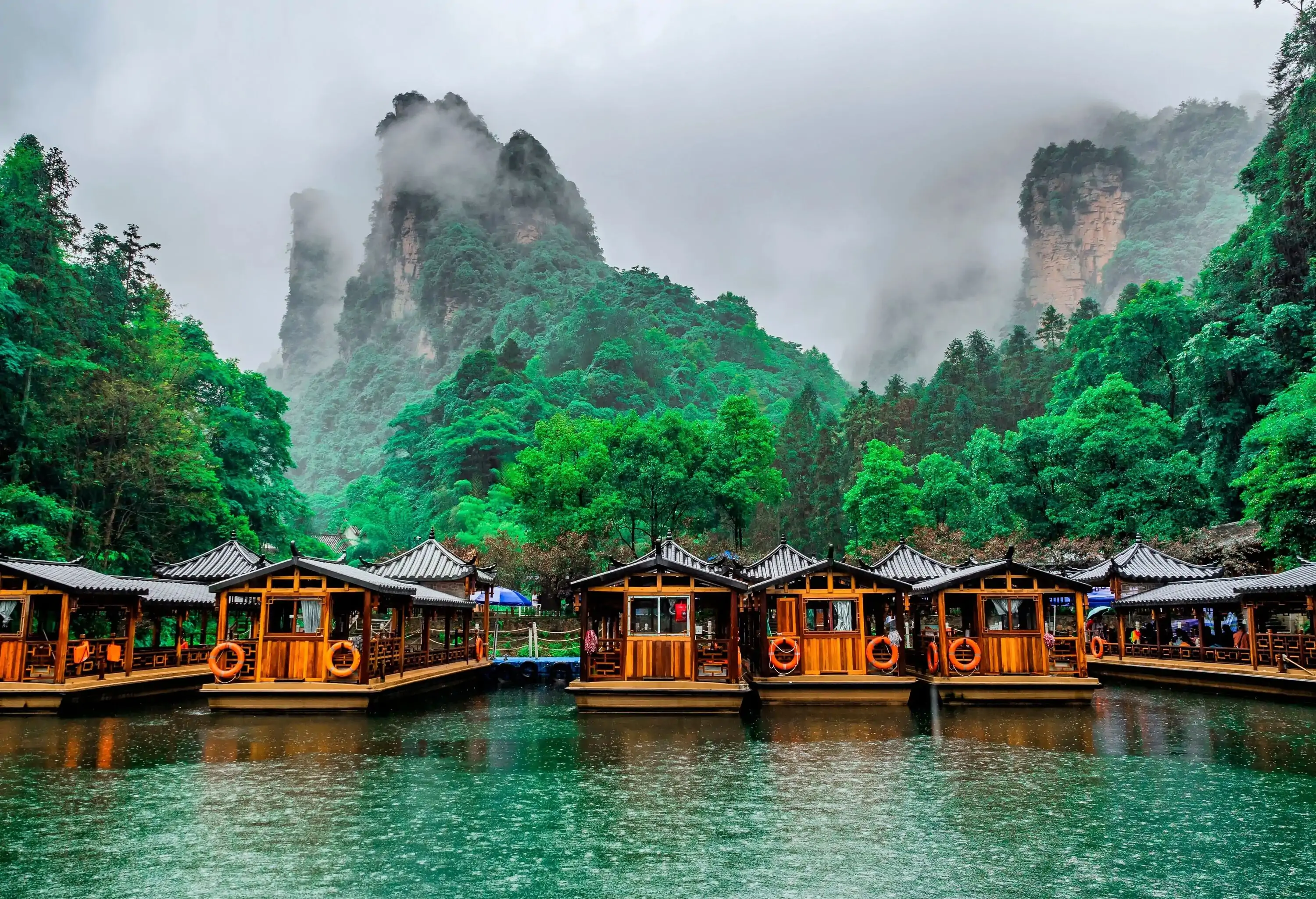Small house-shaped boats docked along a lake surrounded by tall trees and steep mountains covered by fog in Wulingyuan, Zhangjiajie National Forest Park, Hunan Province, China.