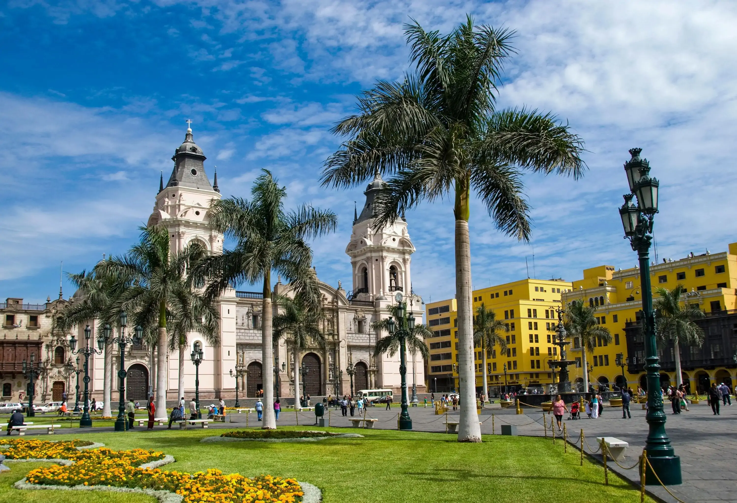 A crowded park with flower gardens and palm trees in front of a cathedral with twin belfries.