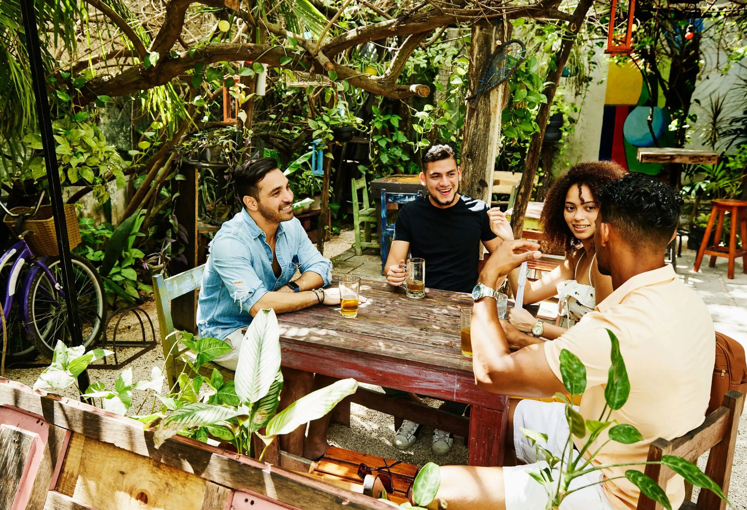 A diverse group of four happy people hanging out in an outdoor cafe surrounded by lush plants.