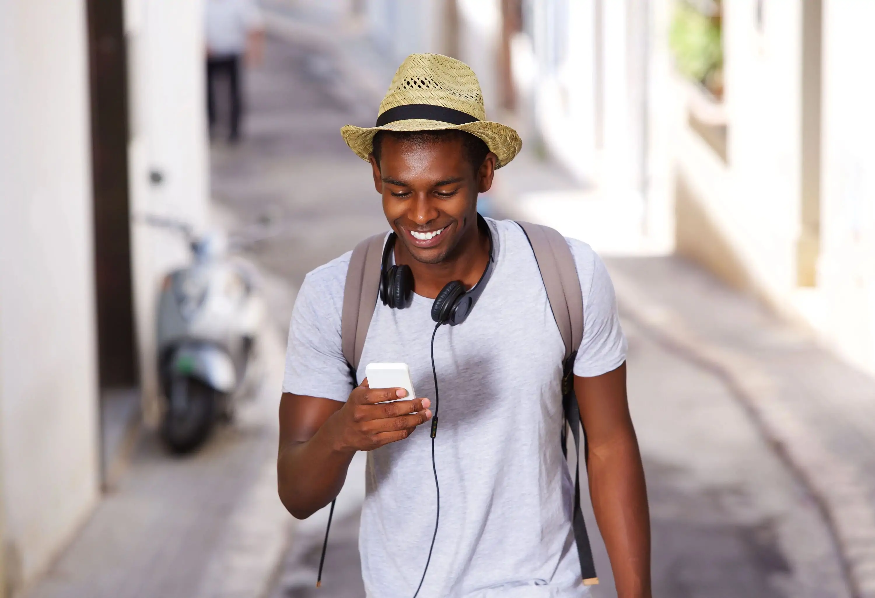 A young man wearing headphones around his neck and staring at his smartphone as he walks through an alley.