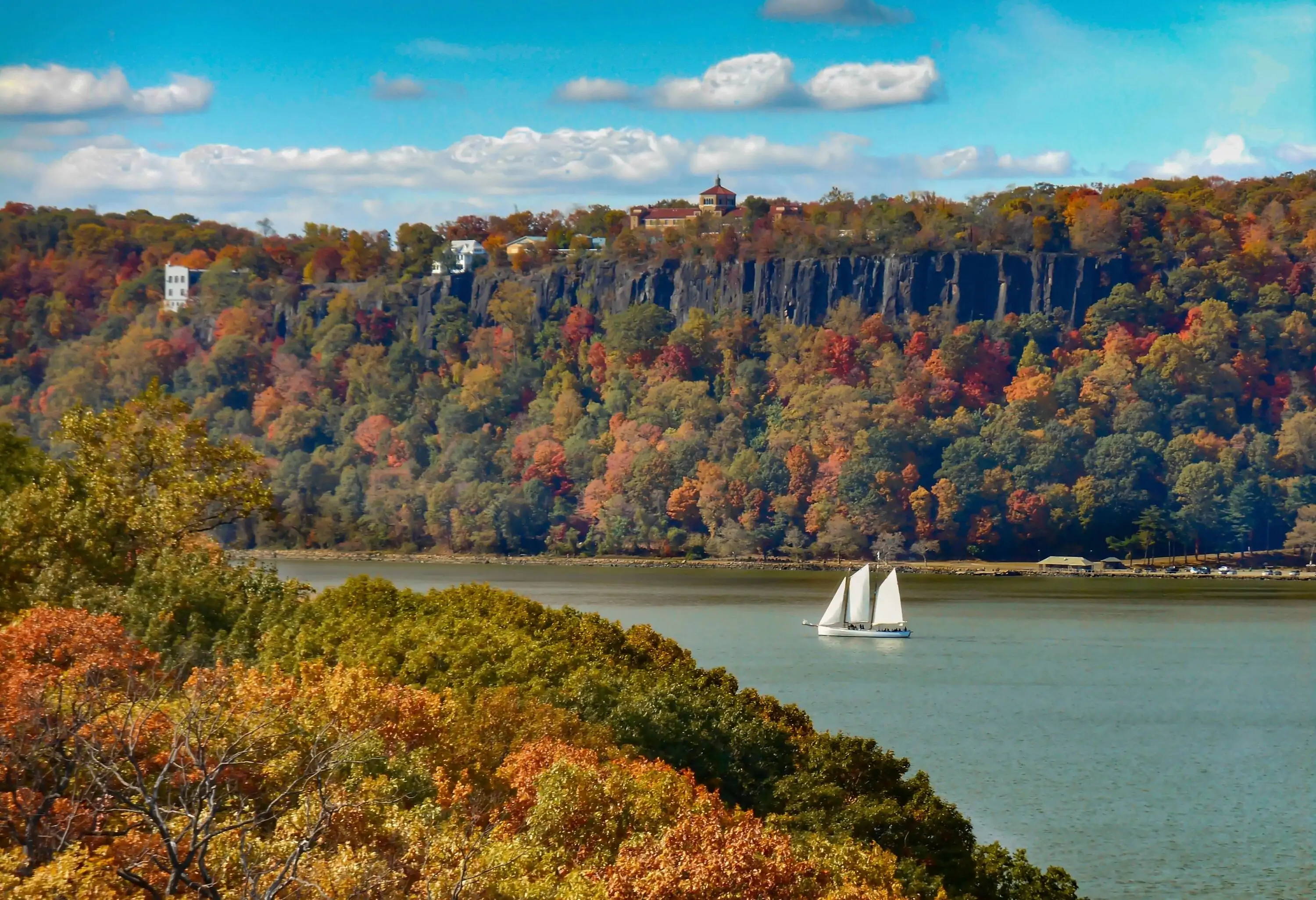 A sailboat on a river between rocky cliffs and thick fall trees.