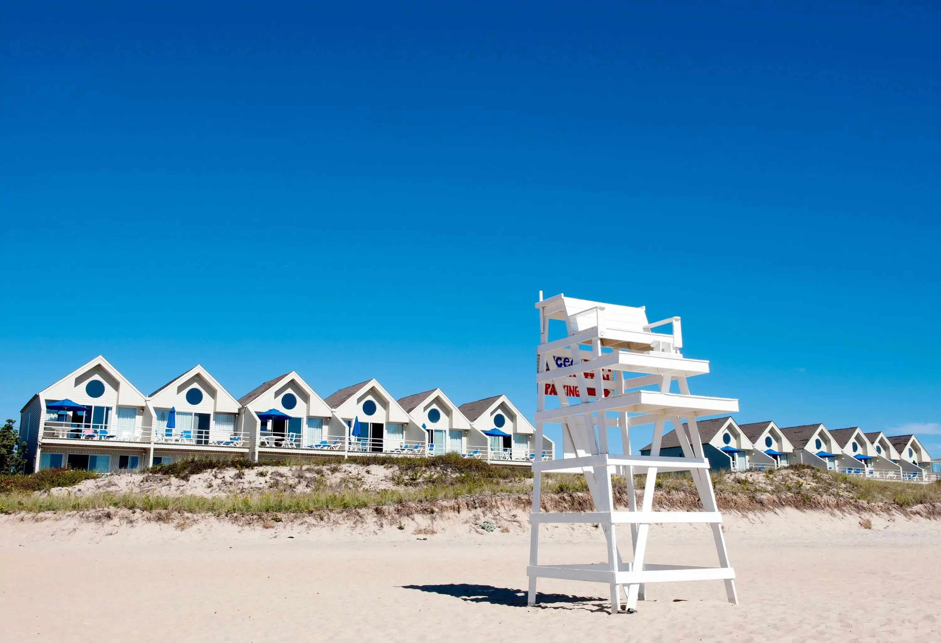 A white lifeguard chair standing on the sand with a row of beach houses in the background.